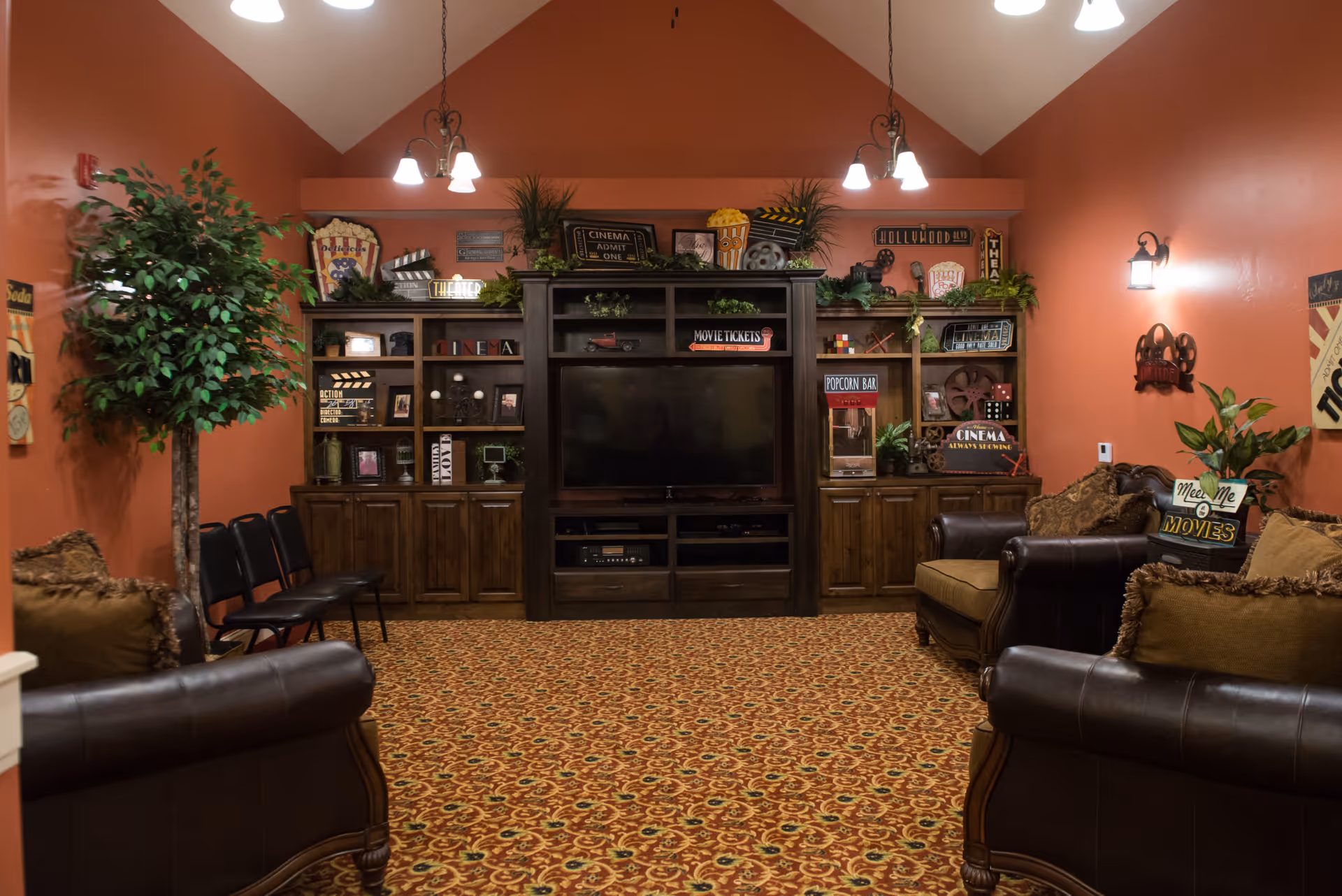A cozy living room with brown leather sofas and armchairs arranged around a large wooden entertainment center with a flat-screen TV. The walls are painted a warm orange color and decorated with movie-themed signs and plants. The carpet has a patterned design, and there are ceiling lights hanging from the vaulted ceiling.