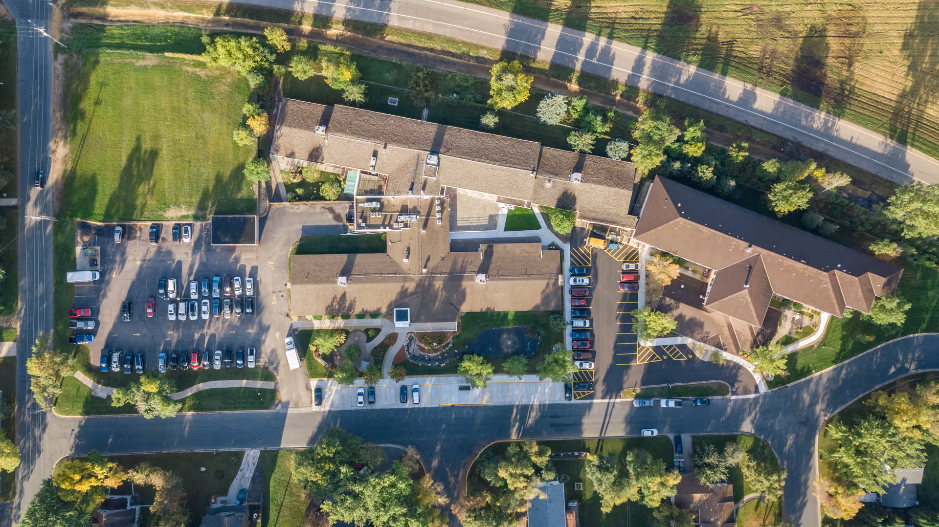Aerial view of Birchwood Healthcare Center SNF showing multiple connected buildings with brown roofs, surrounding parking lots with cars, green lawns, trees, and a road running alongside the facility.