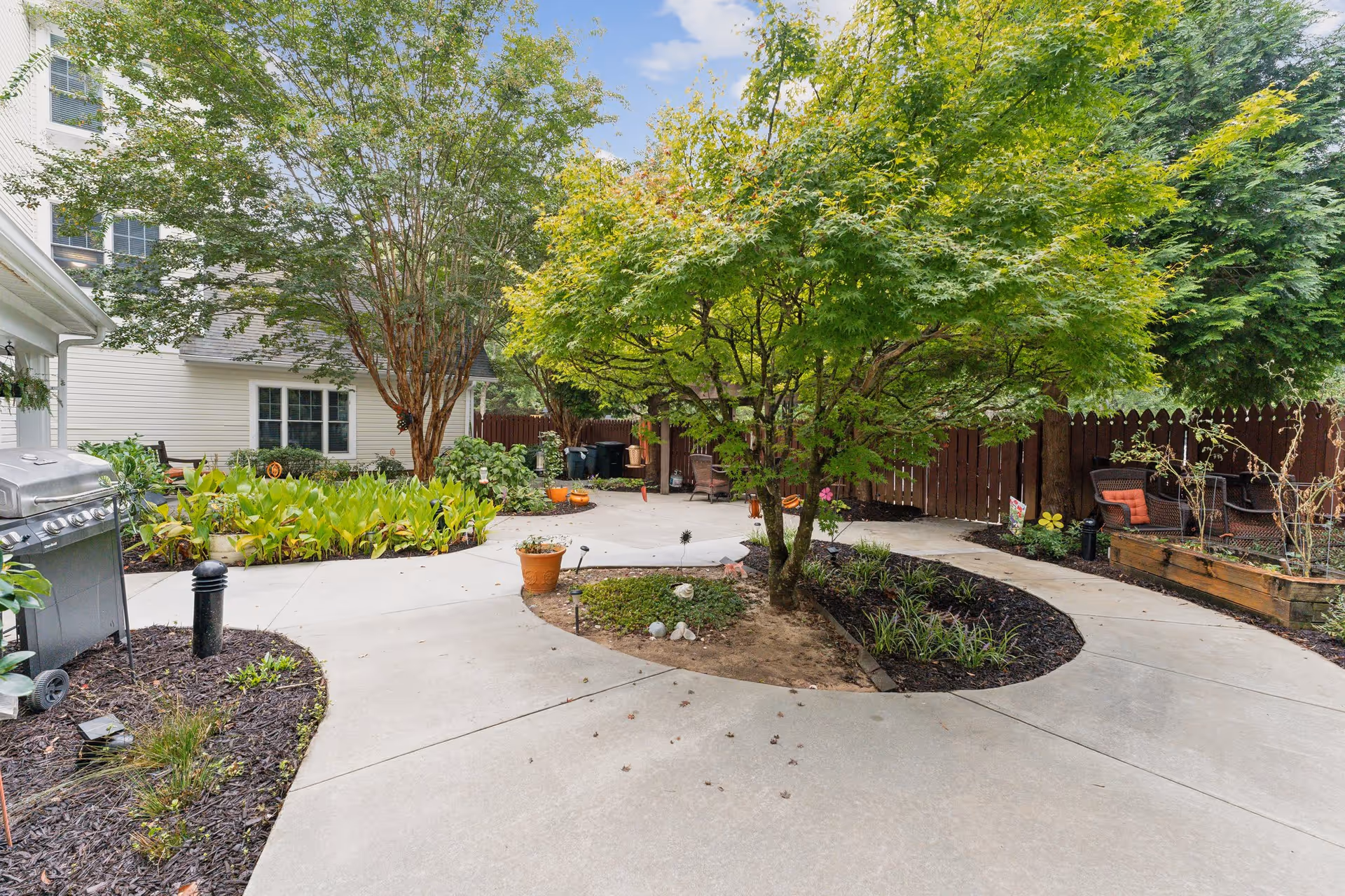 A landscaped outdoor courtyard area with a concrete pathway curving around a small tree and garden beds. There are various plants and shrubs, a barbecue grill on the left side, and outdoor seating with cushions on the right side near a wooden fence. The sky is partly cloudy.