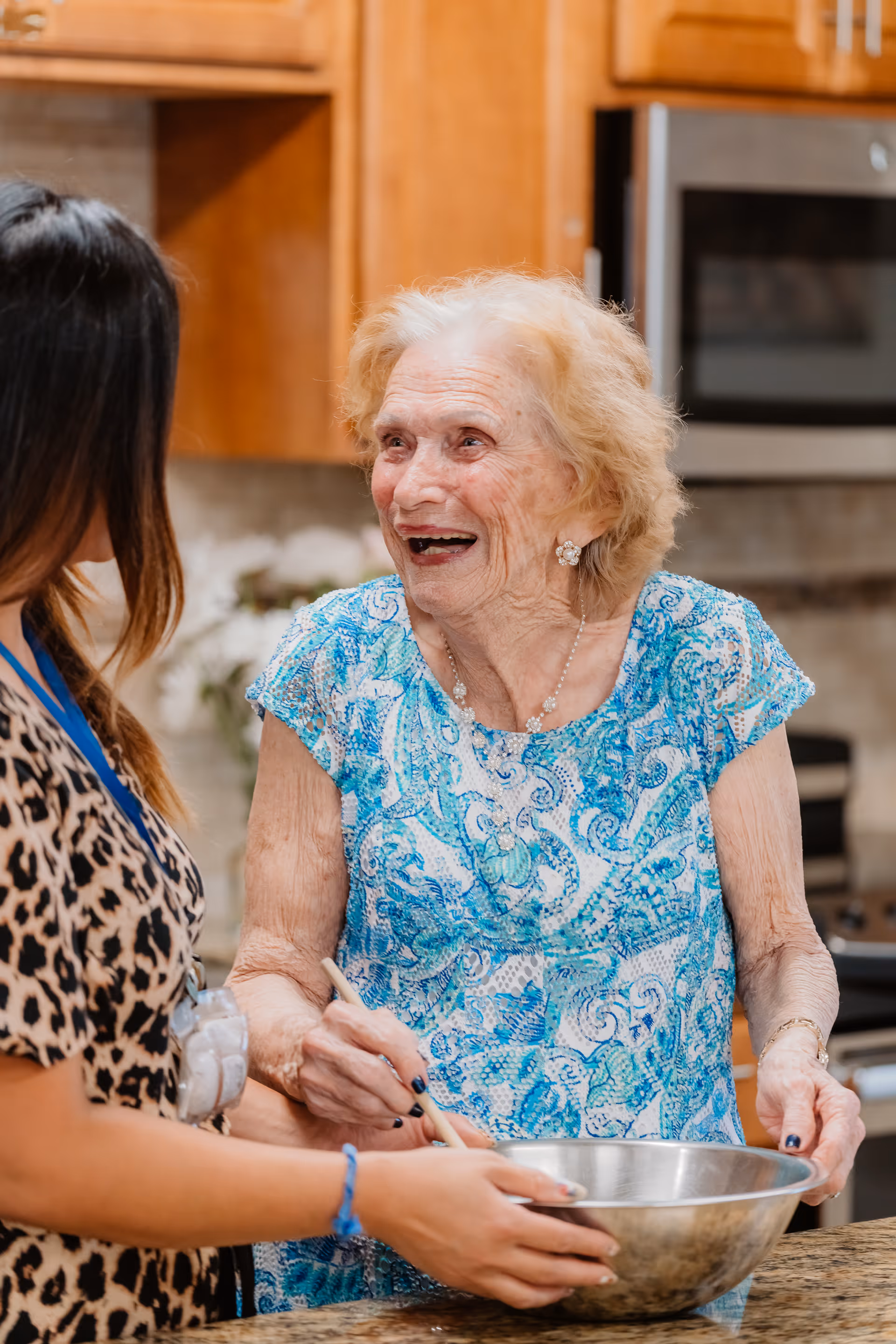 An elderly woman wearing a blue patterned blouse is smiling and stirring a metal mixing bowl while interacting with a younger woman in a kitchen setting with wooden cabinets and a microwave in the background.