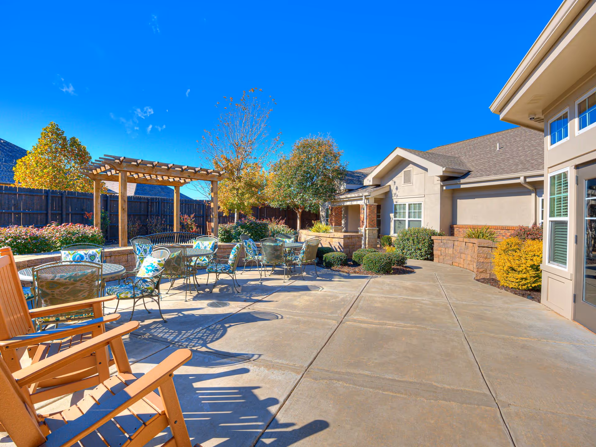 Outdoor patio area at Iris Memory Care Tulsa featuring several metal tables and chairs with cushions, wooden rocking chairs, a pergola, landscaped bushes and trees, and a clear blue sky.