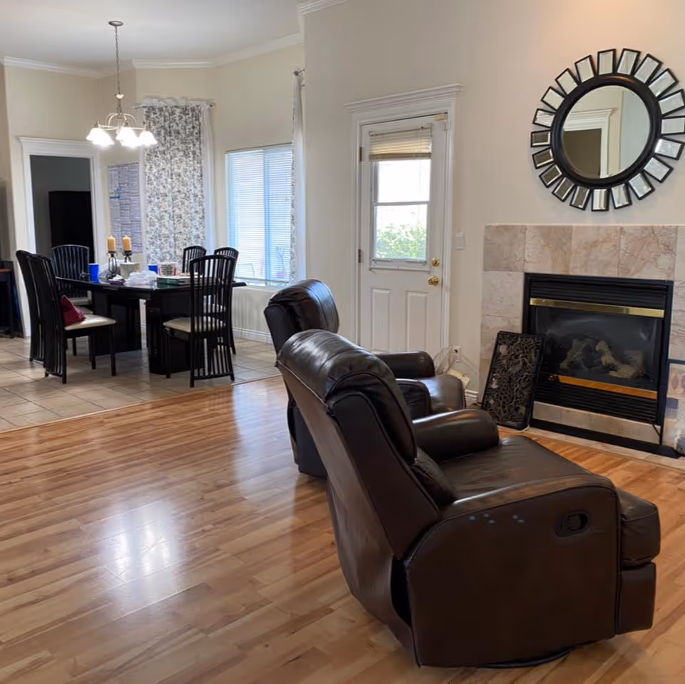 Interior view of a living and dining area in a senior living facility. The foreground shows two brown leather recliners facing a fireplace with a decorative round mirror above it. In the background, there is a dining table with six black chairs, a chandelier overhead, and windows with floral curtains. A white door with a window is next to the fireplace.