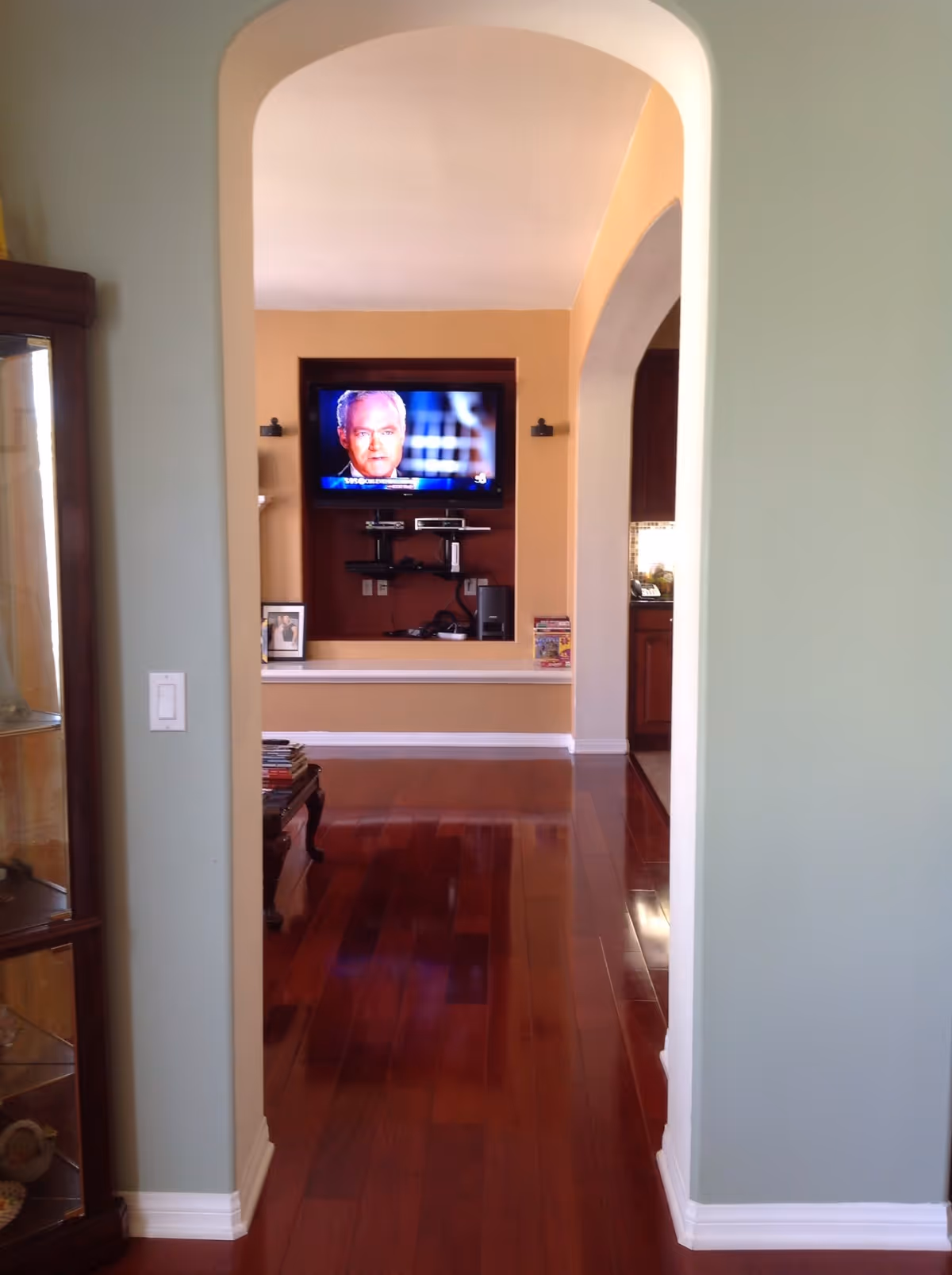 View through an arched doorway into a living room area with polished wooden floors. A flat-screen TV is mounted on a beige wall with a dark brown inset, displaying a man’s face. Below the TV are shelves holding electronic devices. To the left, there is a glass display cabinet with decorative items. To the right, part of a kitchen area with dark wood cabinets is visible.