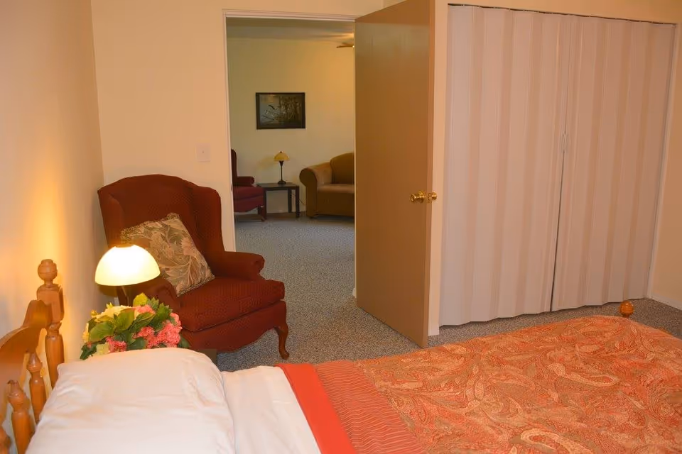 A bedroom with a wooden bed covered with a red patterned blanket and white pillow. Next to the bed is a small lamp and a bouquet of flowers on a nightstand. A red armchair with a floral cushion is placed near the wall. The door is open, revealing a view into a living room area with a sofa, armchair, side table, and a framed picture on the wall.