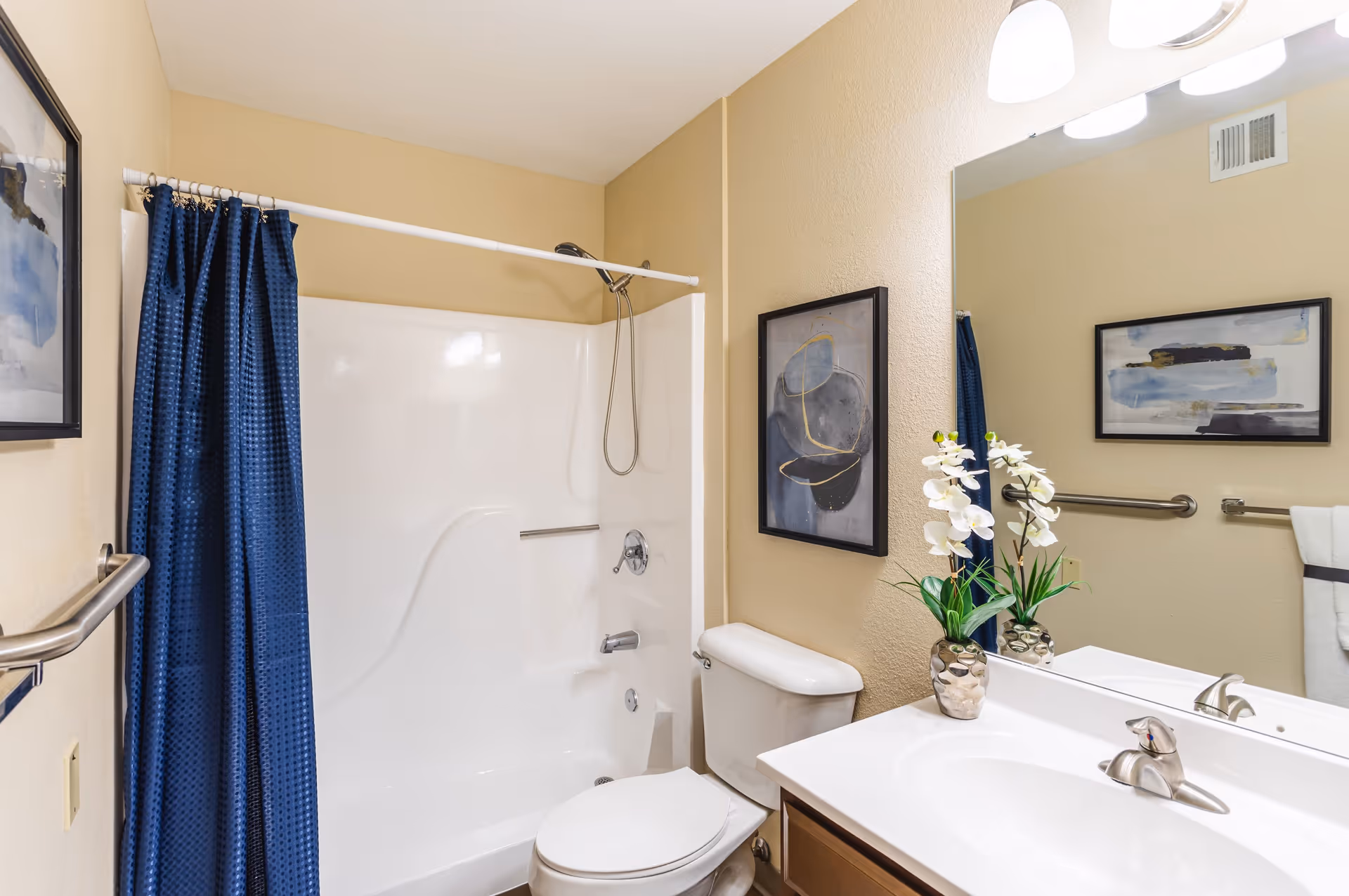 A clean bathroom with a white bathtub and shower combination, a blue shower curtain, a white toilet, a white sink with a silver faucet, a large mirror above the sink, two framed abstract paintings on the walls, and a small vase with white flowers on the sink counter.