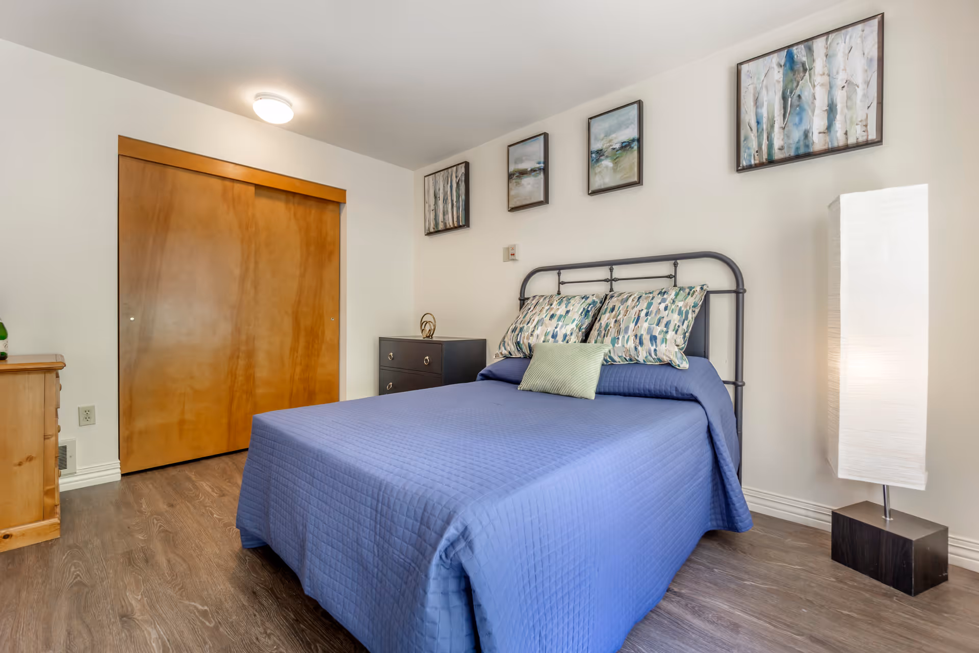 A bedroom with a bed covered in a blue quilt and three decorative pillows. The room has wooden sliding closet doors, a wooden dresser, a black nightstand with a decorative item, and a tall white floor lamp. Four framed artworks hang on the white wall above the bed.