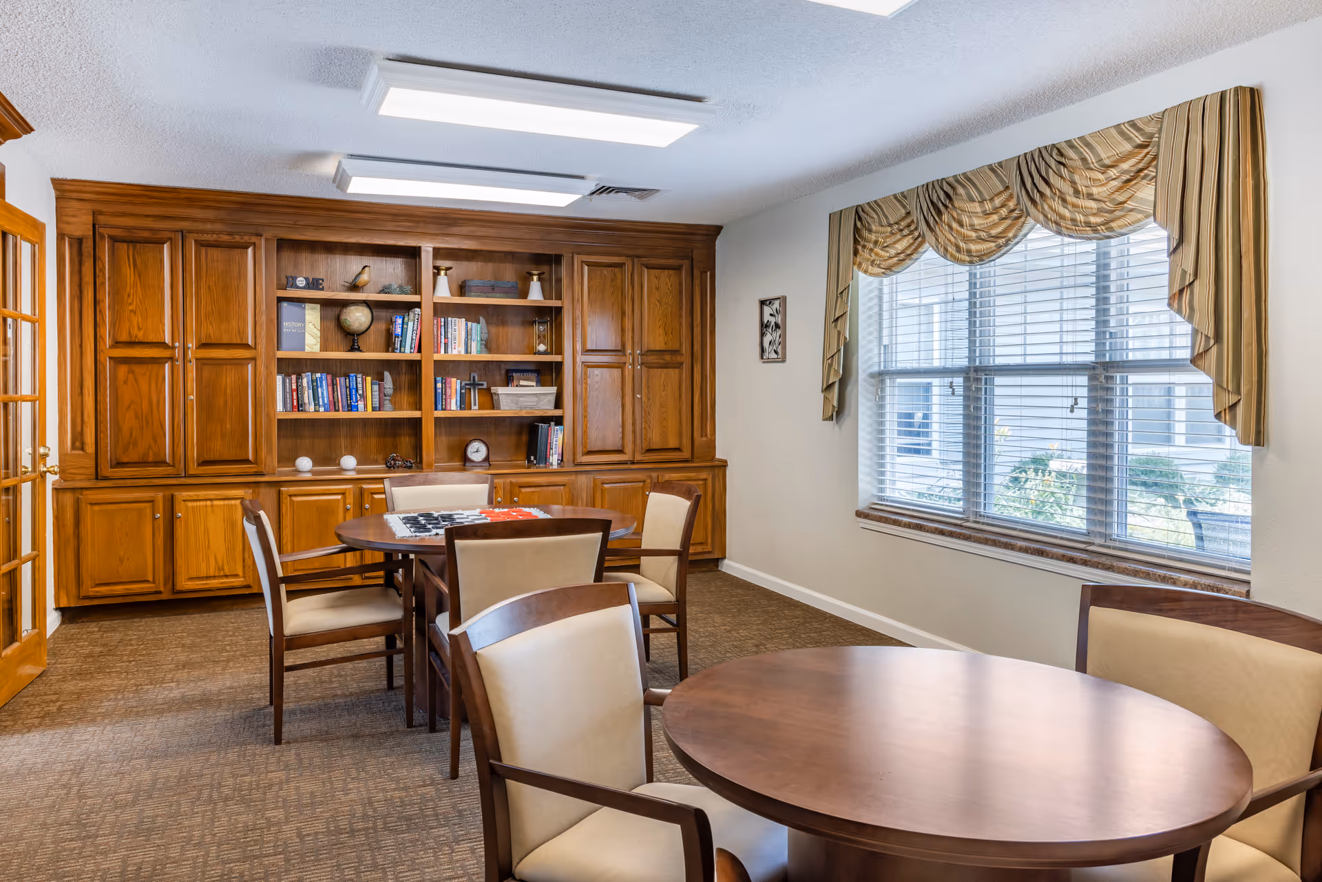 A well-lit room with round wooden tables and cushioned chairs arranged around them. A large wooden built-in bookshelf filled with books and decorative items is against one wall. A window with striped valance curtains lets in natural light, and the carpeted floor complements the warm wood tones of the furniture.