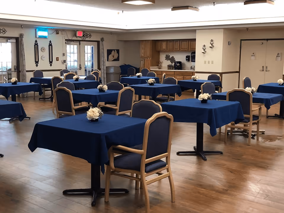 Spacious dining room with tables covered in blue tablecloths and chairs arranged around them, each table topped with a small white floral centerpiece.
