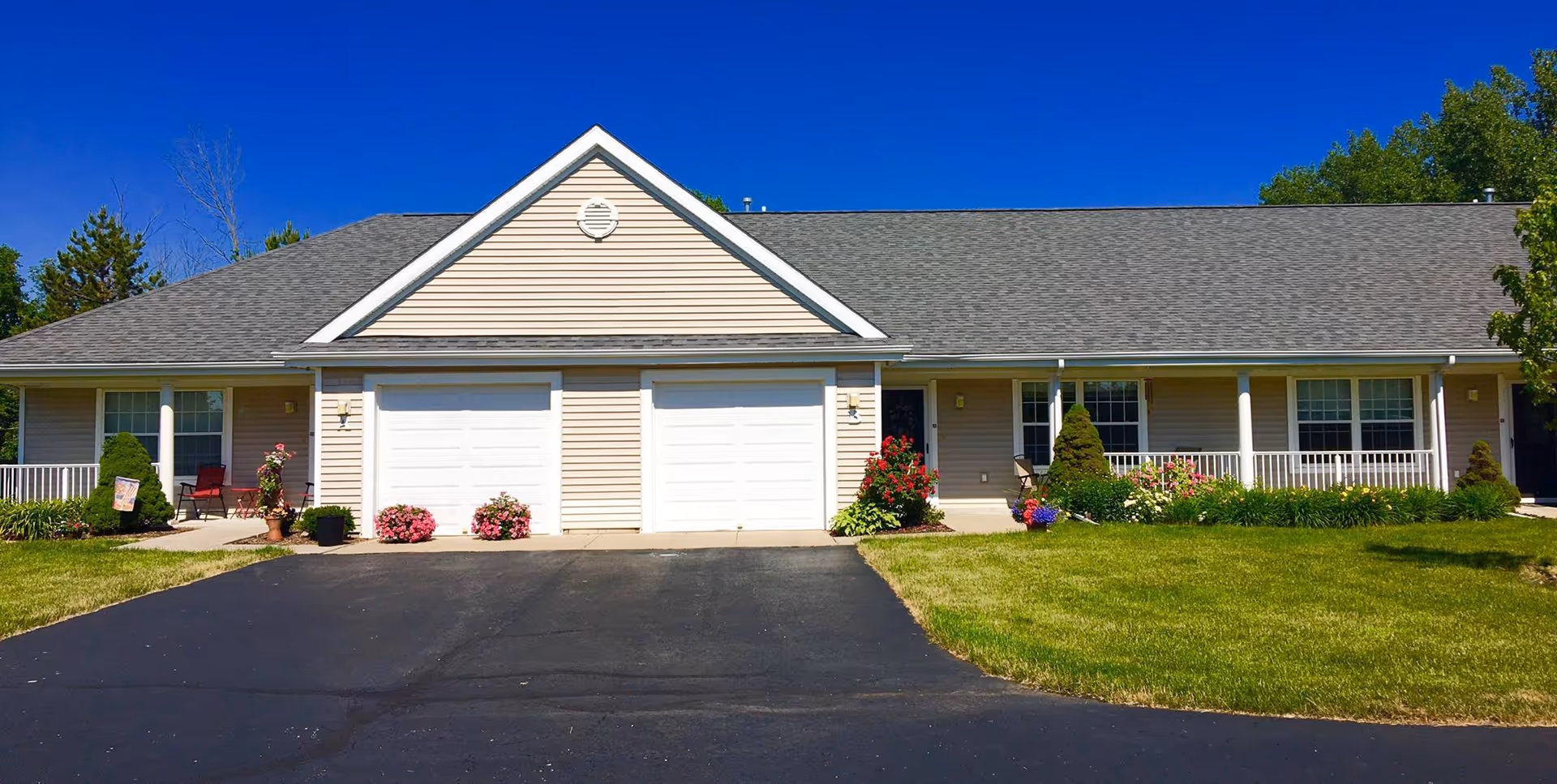 Single-story building front with two white garage doors, covered porches, flower beds and a mowed lawn under a clear blue sky.