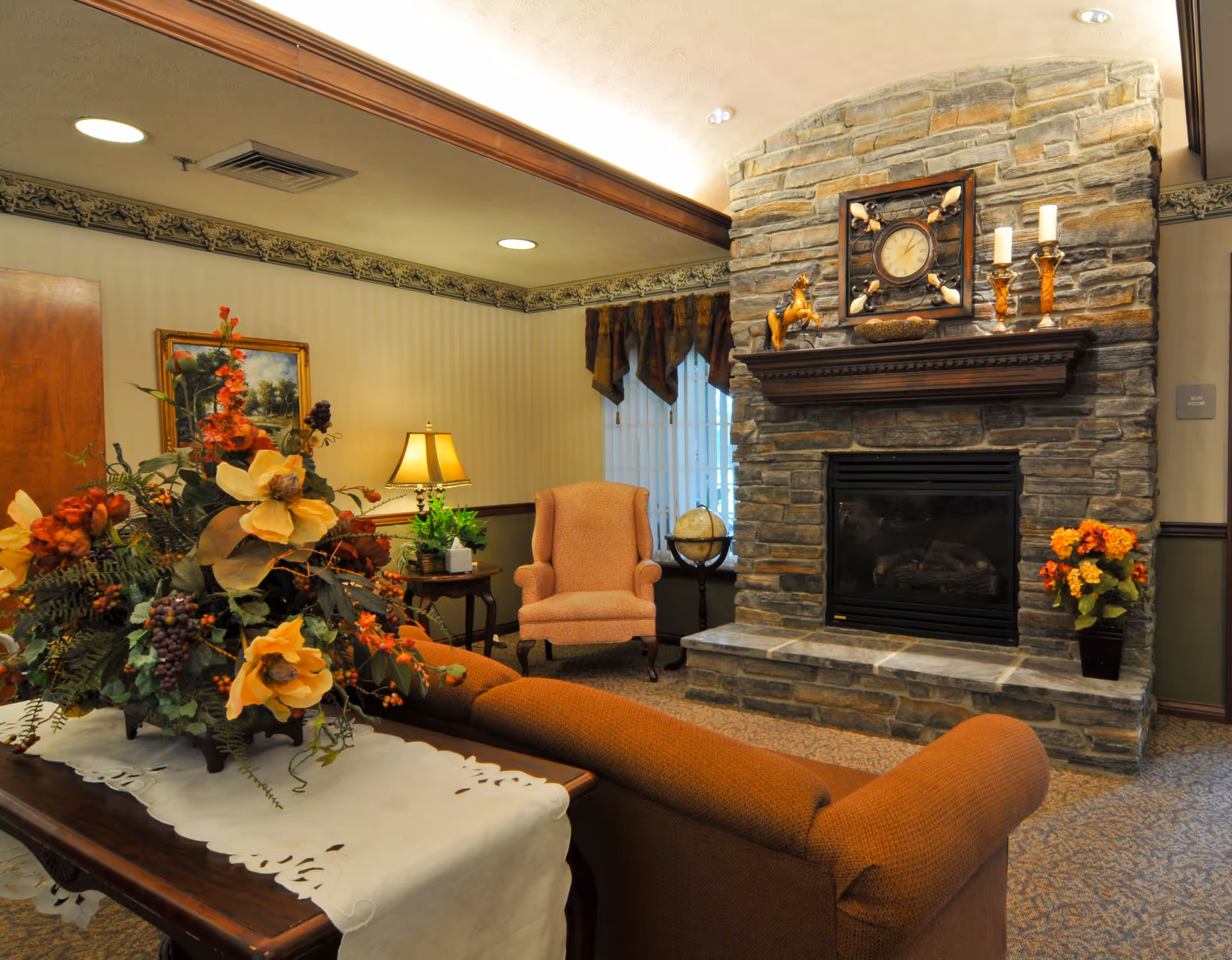 A cozy living room area featuring a stone fireplace with a clock and decorative items on the mantel. In front of the fireplace is a comfortable armchair and a side table with a lamp and plants. A sofa faces the fireplace, and a wooden table with a white runner and a large floral arrangement is in the foreground. The room has warm lighting and traditional decor.