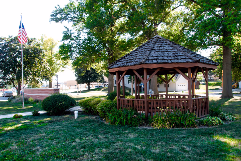 A wooden gazebo with a shingled roof surrounded by greenery and bushes in a grassy outdoor area. An American flag on a flagpole is visible to the left, with trees and buildings in the background under a clear sky.