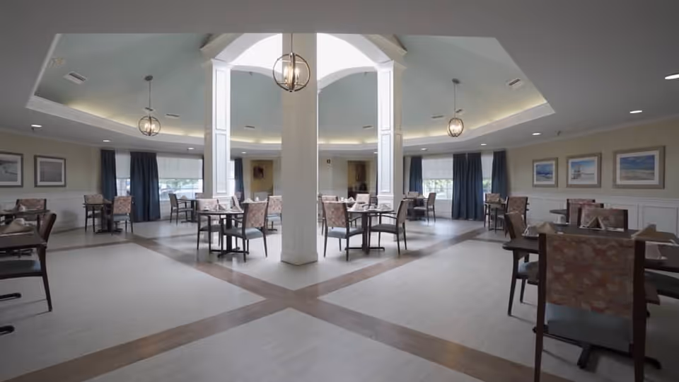 A spacious dining room with multiple tables and chairs arranged neatly. The room features a high ceiling with modern hanging light fixtures, large windows with dark curtains, and framed artwork on the walls. The floor has a light-colored tile with a darker tile pattern creating a cross shape in the center.