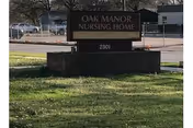 A brick front sign on a grassy lawn that reads "OAK MANOR NURSING HOME" with the number "2901" on the base and a parking lot and buildings behind it.