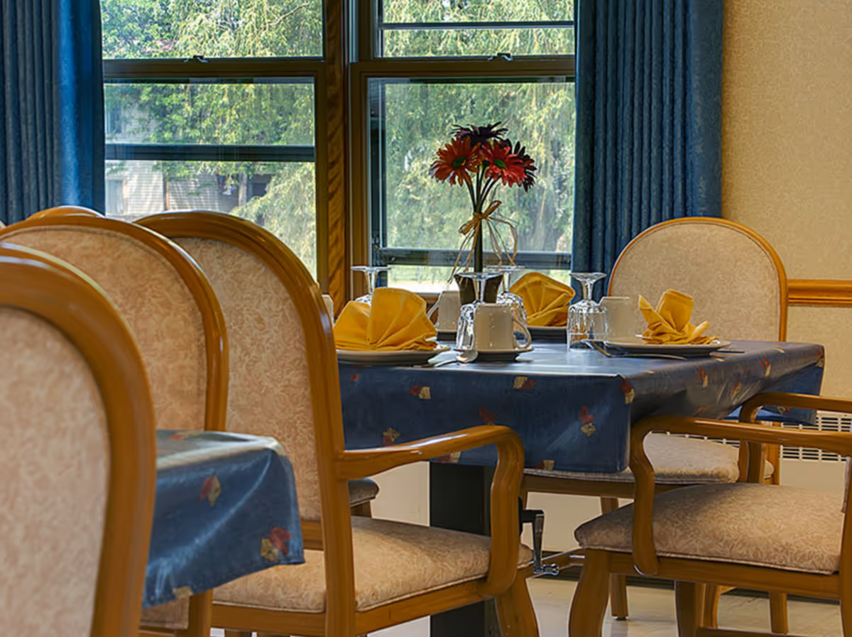 Dining area with a table covered in a blue tablecloth set with yellow folded napkins, white cups, and glasses. There are wooden chairs with cushioned seats and backs around the table. A vase with red flowers is placed in the center of the table. Large windows with blue curtains provide a view of green trees outside.