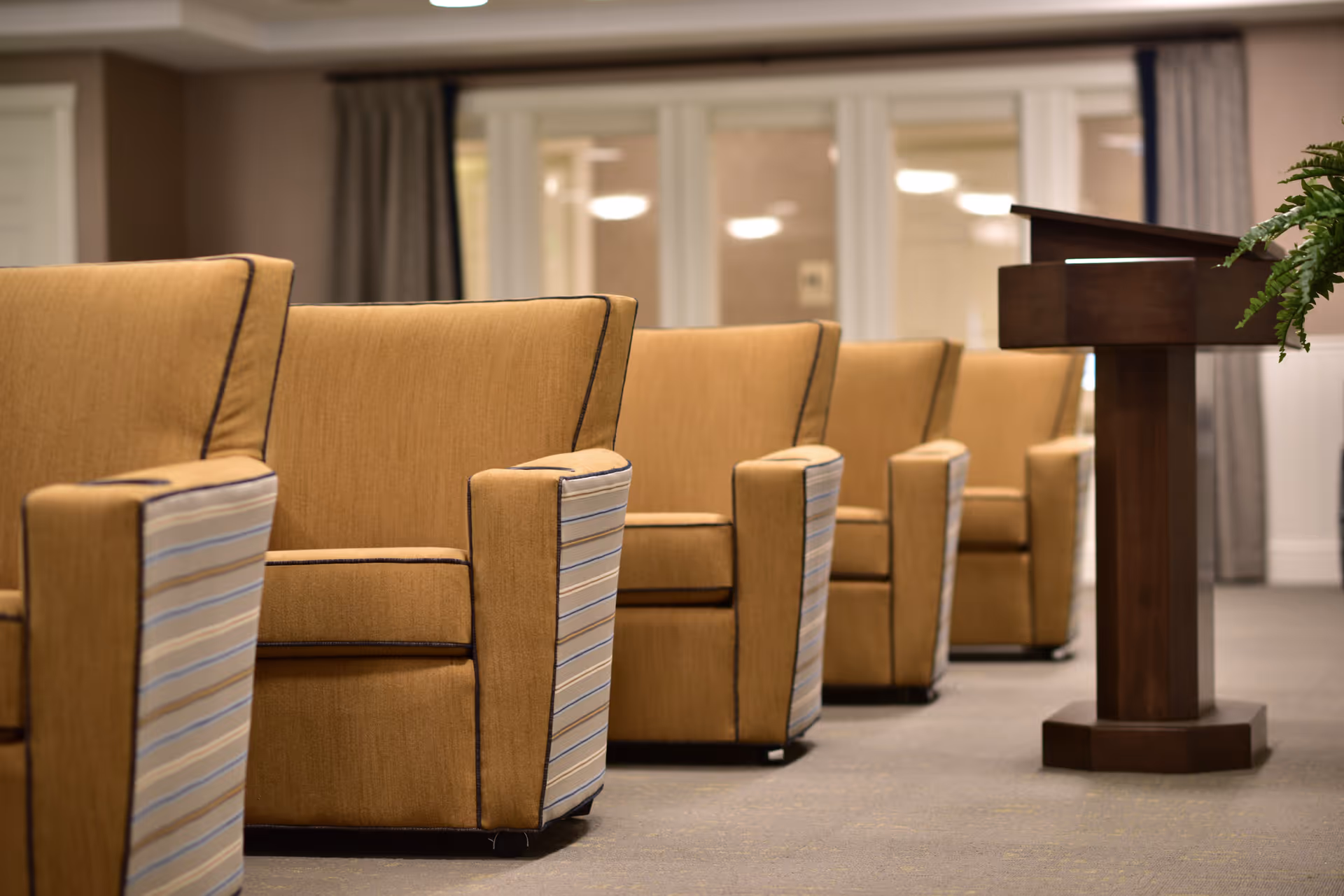 A row of beige upholstered armchairs with striped sides arranged in a room facing a wooden podium with a plant on the right side, with windows and curtains in the background.
