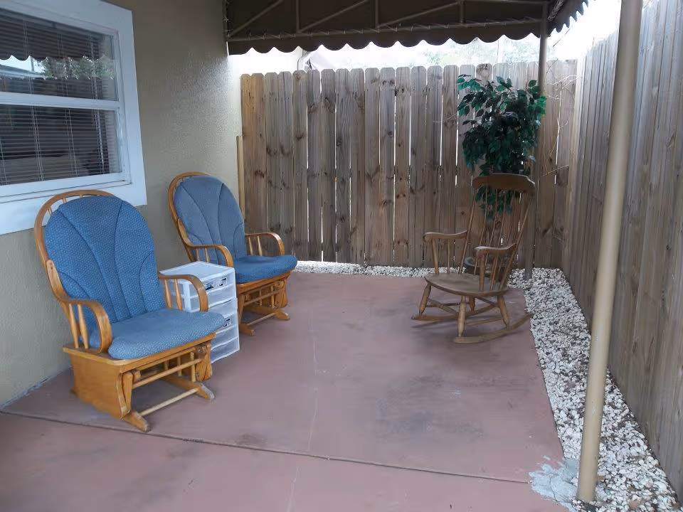 Covered outdoor patio with two blue-upholstered wooden glider chairs, a wooden rocking chair, a small plastic drawer unit, and a potted plant by a wooden fence.