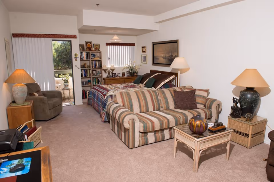 A cozy retirement living space featuring a striped sofa with a brown pillow, a wooden coffee table with decorative items, a bed with a colorful quilt, a bookshelf filled with books and decorations, two table lamps, a comfortable armchair, and a sliding glass door leading to an outdoor patio area.