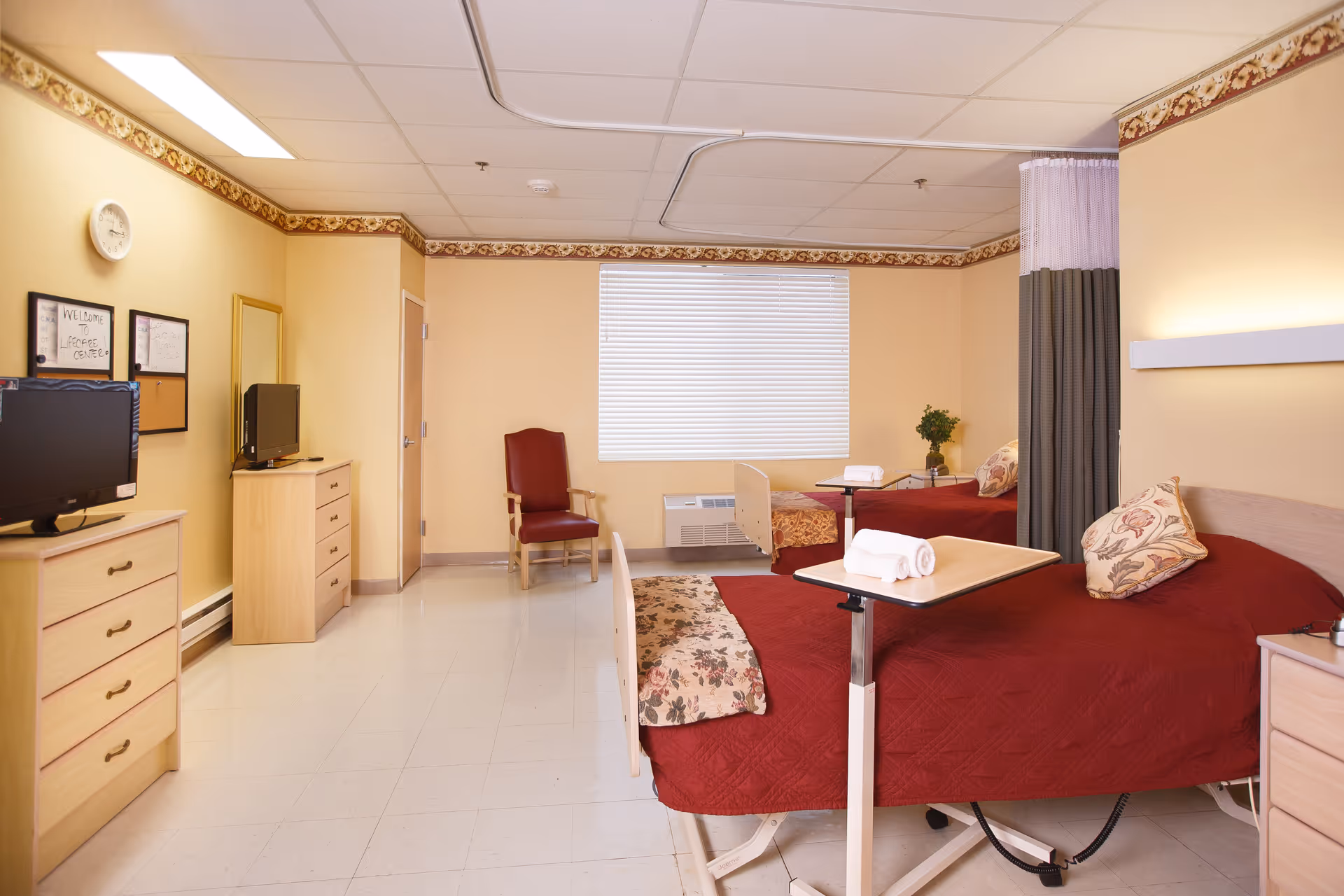 A bright senior care bedroom with two beds dressed in red bedspreads, bedside tables, a TV on a dresser, and a chair.