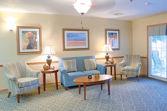 A cozy living room with a blue sofa and two striped armchairs arranged around a wooden coffee table. Two side tables with matching lamps flank the sofa. Three framed paintings hang on the beige walls, and a large window with blinds lets in natural light.
