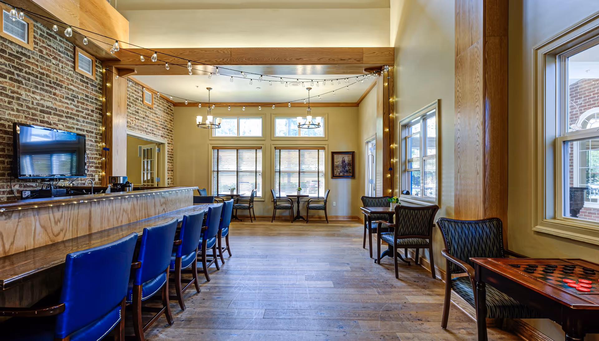 A spacious common area with a long wooden bar counter lined with blue chairs on the left side. The room features exposed brick walls, large windows with blinds, and string lights hanging from the ceiling. On the right side, there are small tables with chairs, one of which has a checkers board on it. The floor is wooden, and the room is well-lit with natural light coming through the windows.