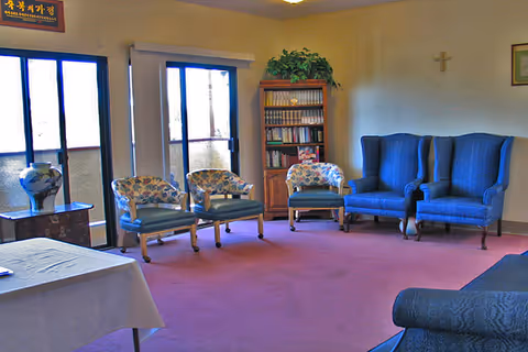 A sitting room with a purple carpet featuring three floral upholstered chairs and two blue wingback chairs arranged along the walls. There is a wooden bookshelf filled with books and a green plant on top. A table with a white tablecloth is partially visible in the foreground. Large windows and a sliding glass door let in natural light. The walls are light-colored with a small cross and framed pictures hanging.