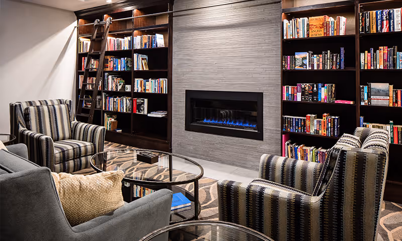 Cozy living room area with two striped armchairs and a gray sofa surrounding a glass coffee table. Behind the seating is a modern electric fireplace set in a tiled wall, flanked by dark wooden bookshelves filled with books and a rolling ladder for access.