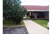 Outdoor view of a pathway leading to the entrance of a single-story building with a red roof and large windows. There is a tree and grass on the left side of the pathway and a small patch of grass on the right side.