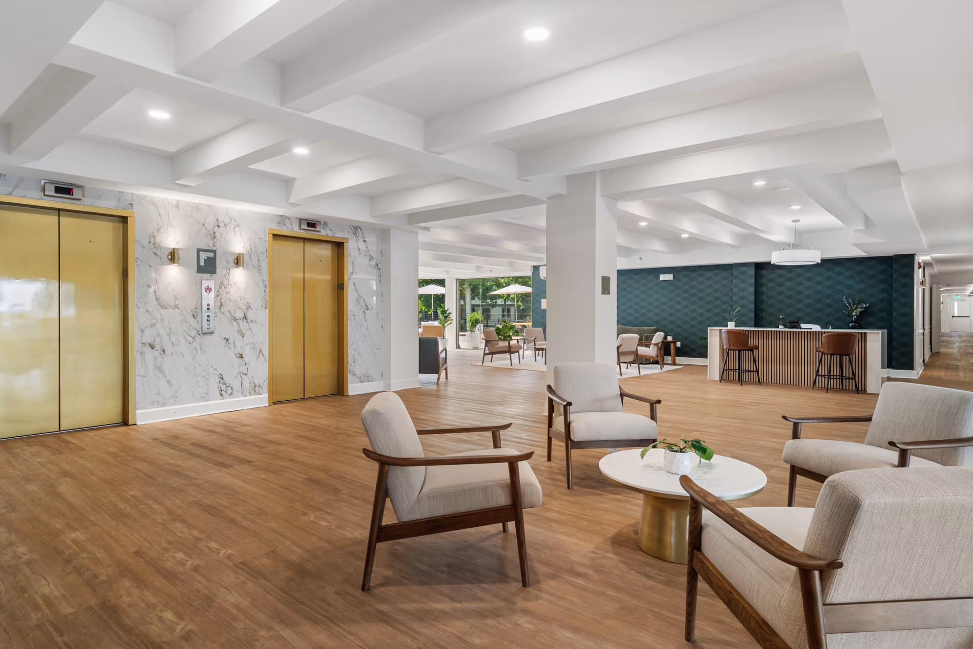 A spacious and modern lobby area with wooden flooring, four beige cushioned chairs arranged around a round white table with a small plant on it. To the left, there are two gold-colored elevator doors set in a white marble wall. In the background, there is a reception desk with two brown chairs and a dark green textured wall behind it. Large windows and glass doors reveal an outdoor patio area with umbrellas and seating.