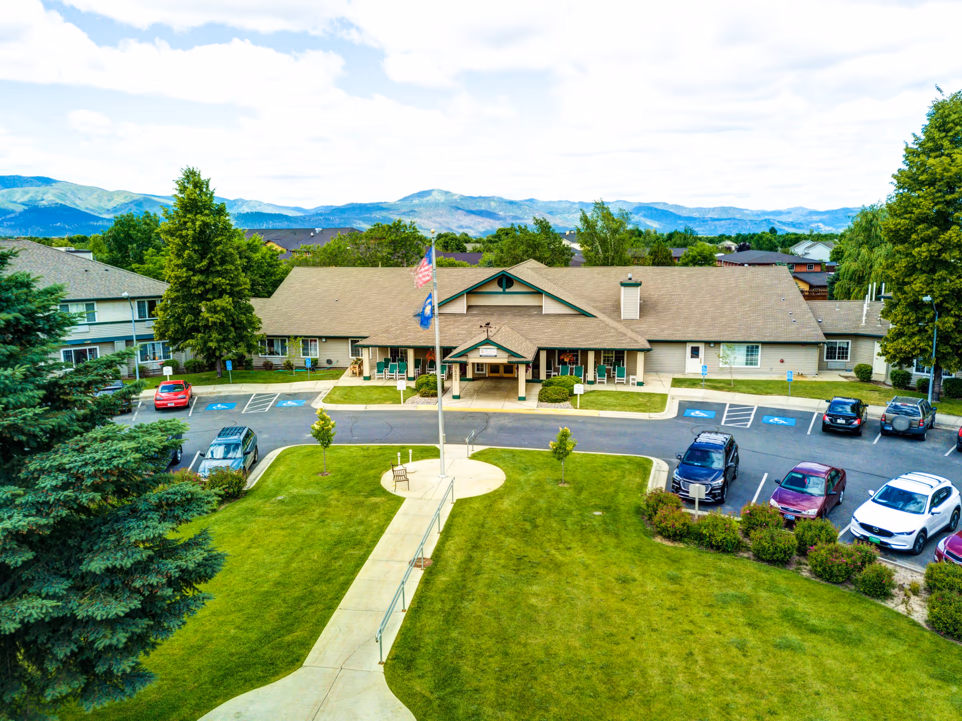 Front exterior view of Missoula Valley Senior & Assisted Living facility showing a single-story building with a peaked roof, a covered entrance with chairs, a flagpole with two flags, a parking lot with several cars, and a well-maintained lawn with trees and shrubs. Mountains are visible in the background under a partly cloudy sky.