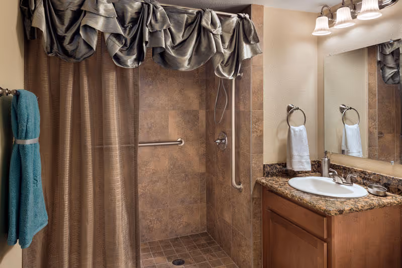 A bathroom with a walk-in shower featuring brown tiled walls and floor, a silver grab bar, and a shower curtain with a decorative valance. There is a wooden vanity with a granite countertop, a white sink, a soap dispenser, and a hand towel hanging on a ring. A large mirror and three light fixtures are mounted above the sink.