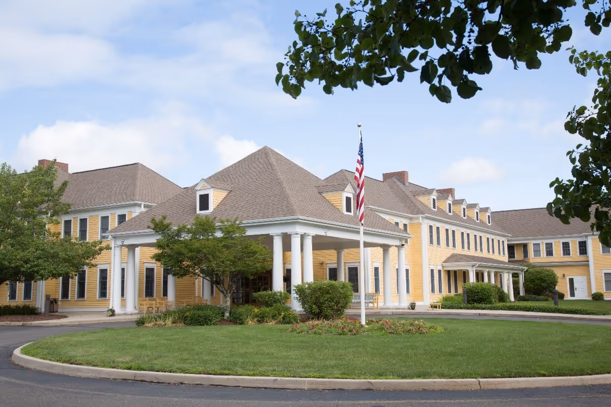 Exterior view of a large yellow senior living facility building with white columns at the entrance, a circular driveway, green lawn, trees, and an American flag on a flagpole in front.