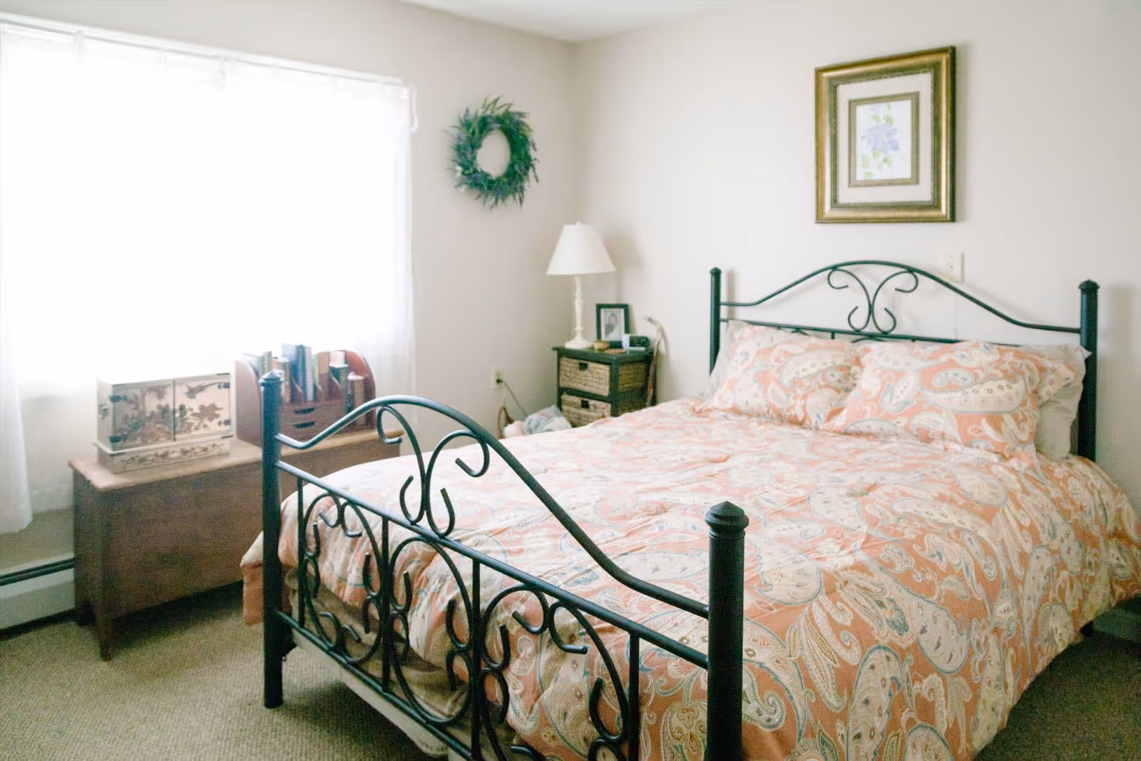 A cozy bedroom with a black metal bed frame and a bed covered in a peach and white patterned comforter. There is a wooden chest near a window with sheer white curtains, a small nightstand with a lamp and picture frame, a green wreath hanging on the wall, and a framed floral picture above the bed.