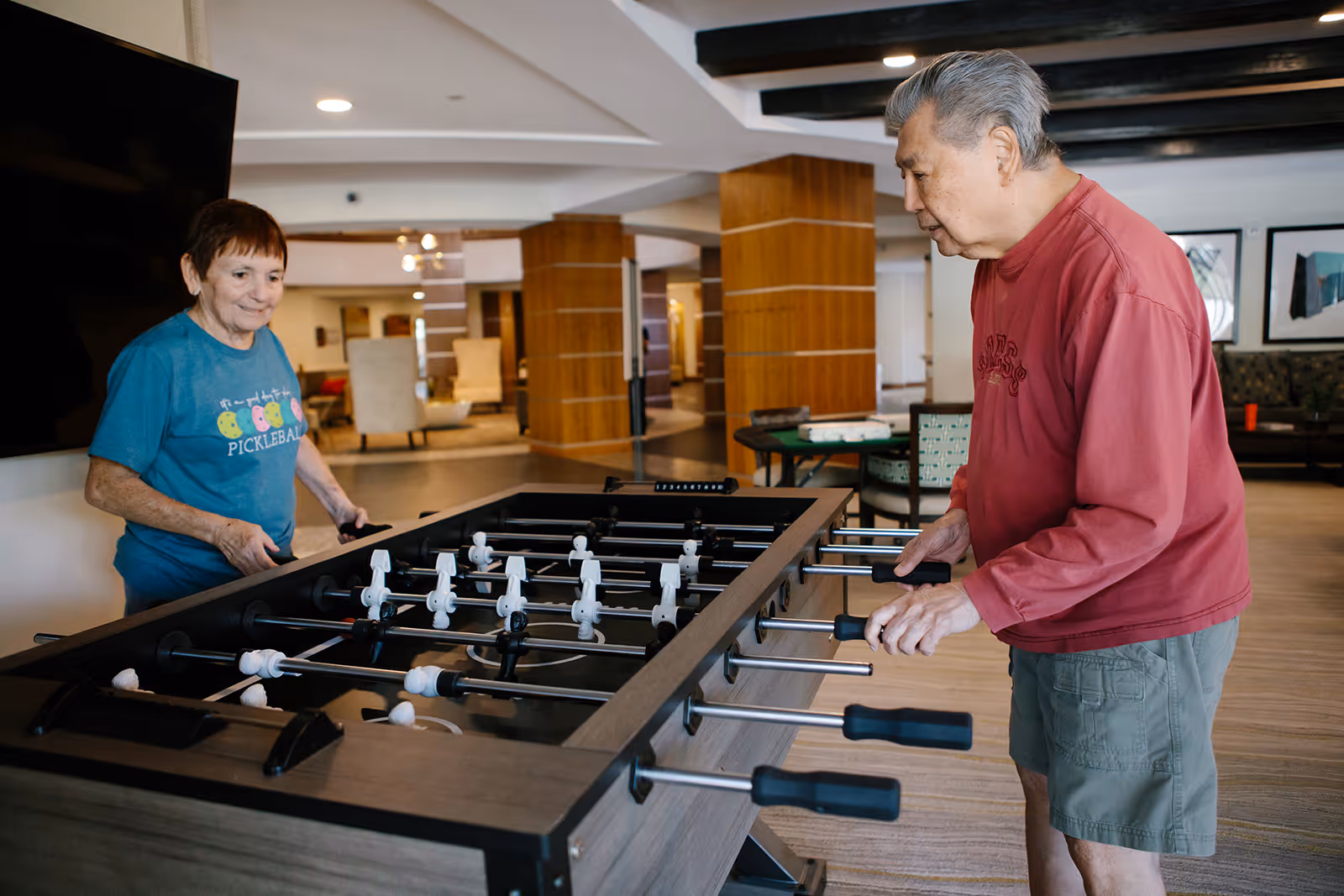 Two elderly people playing foosball in a spacious, well-lit common area with wooden pillars and comfortable seating in the background.