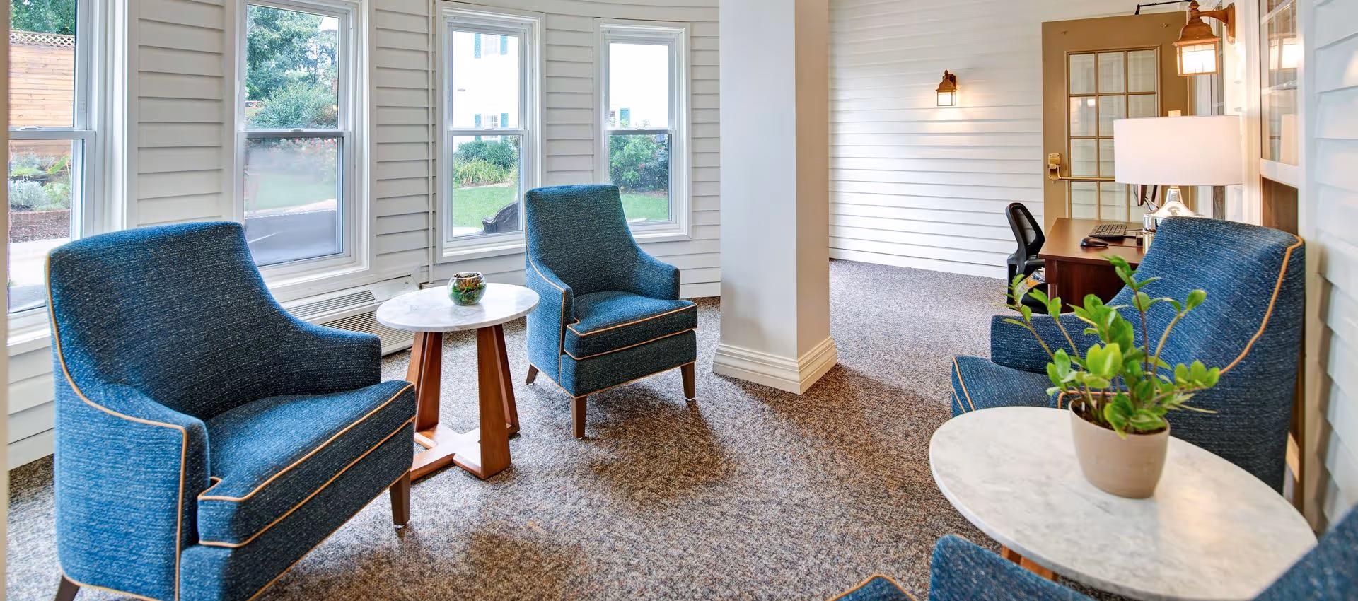 Sunny sitting area with blue upholstered armchairs, round side tables, windows and a desk with a lamp and plant.