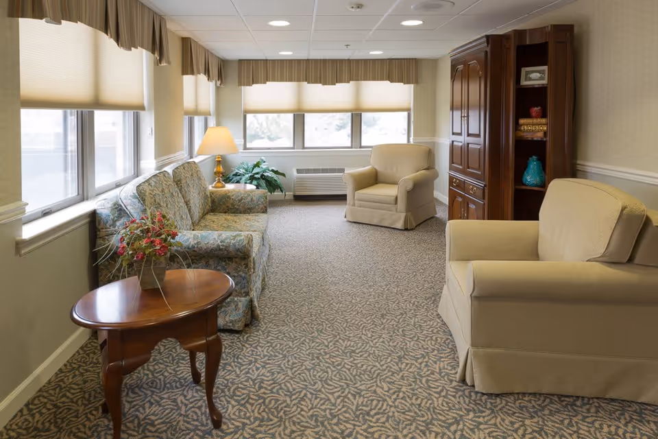 Sunlit sitting room with a patterned sofa, two beige armchairs, wooden side tables and a cabinet beneath multiple windows.
