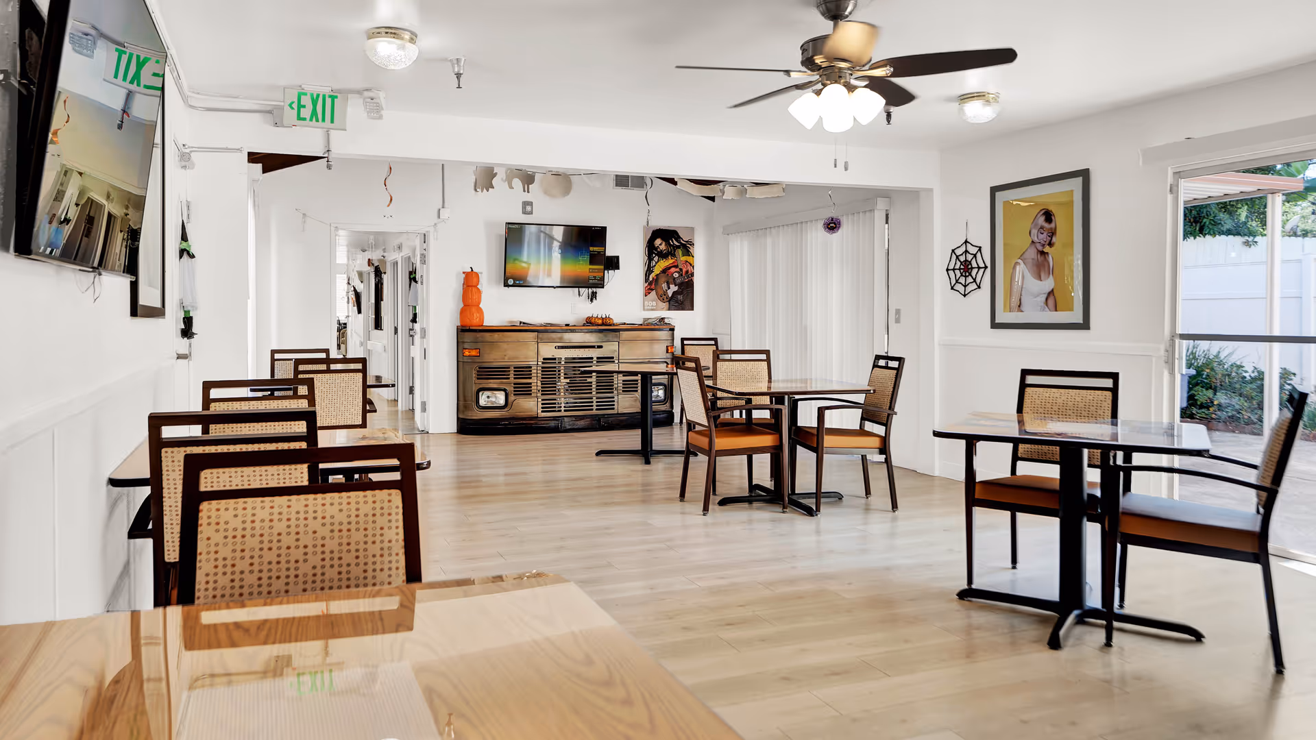 A bright dining room with several tables and chairs arranged neatly. The room has light wooden flooring, white walls, a ceiling fan with lights, and a wall-mounted TV. Decorations include a framed picture, a spider web decoration, and a pumpkin stack on a sideboard. Large windows and sliding glass doors allow natural light to enter, showing some greenery outside.
