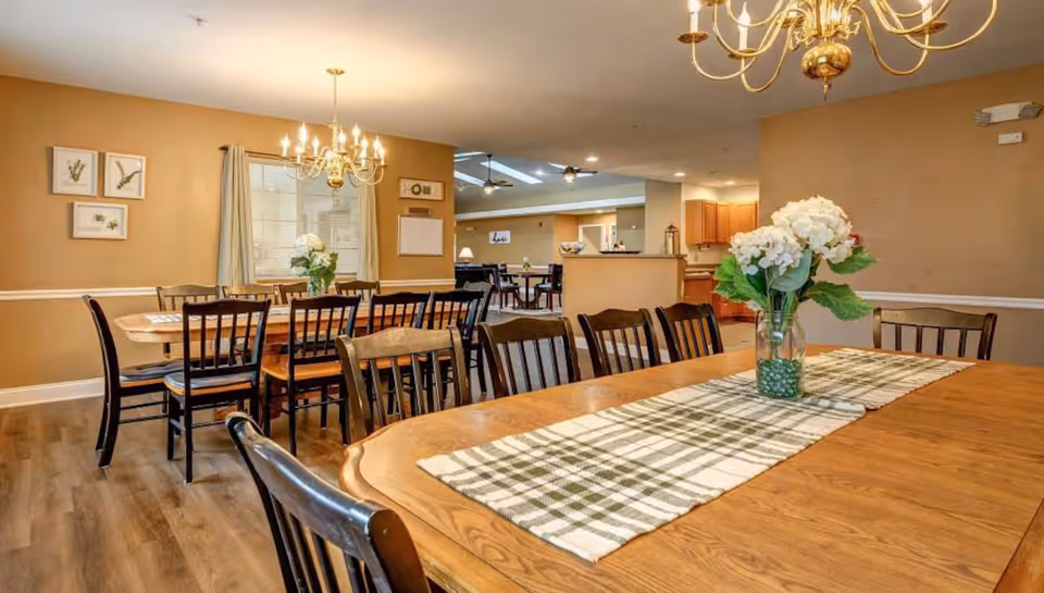 A warm dining room with wooden tables, chairs, chandeliers, and a vase of flowers.