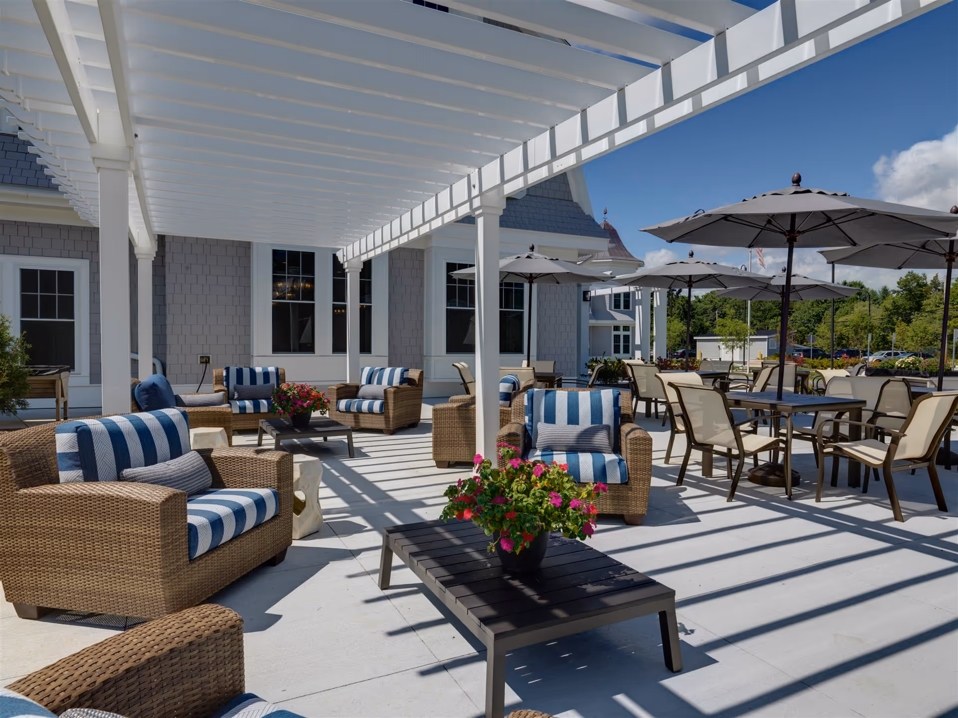 Outdoor patio area at The Residence at Penniman Hill featuring wicker armchairs with blue and white striped cushions, tables with umbrellas, and a white pergola casting shadows on the ground. There are potted flowers on the tables and a clear blue sky in the background.