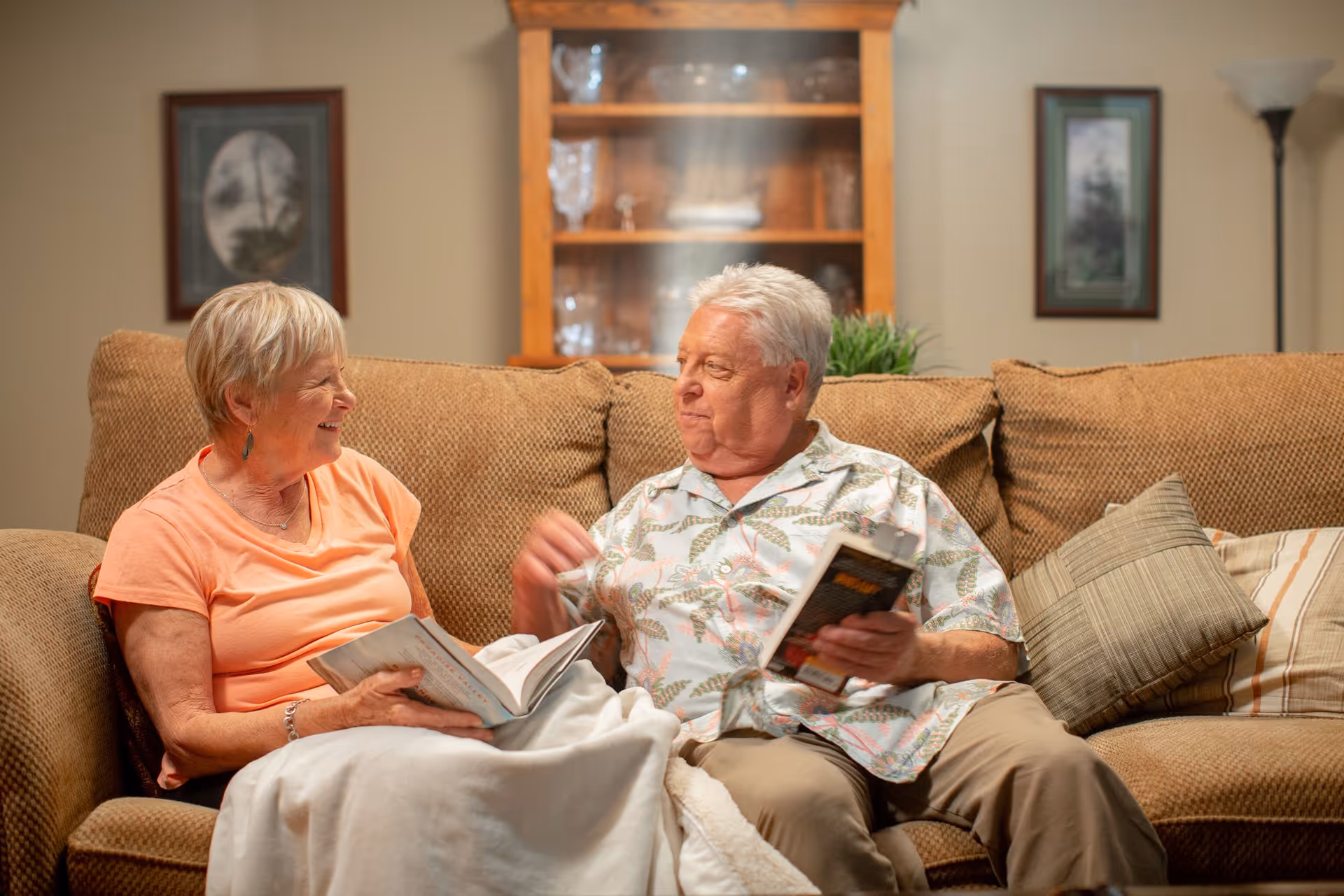 Two older adults sitting on a couch in a living room, smiling and holding books.
