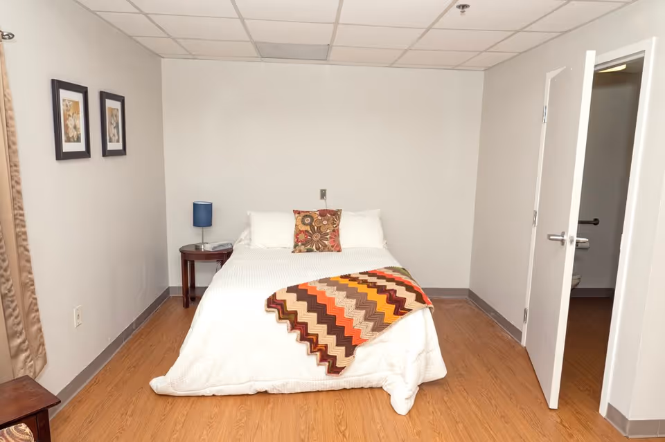 A simple bedroom with a bed covered in a white bedspread and a colorful zigzag patterned blanket. There is a decorative pillow on the bed. A small wooden nightstand with a blue lamp and a phone sits to the left of the bed. Two framed pictures hang on the left wall. The floor is wood, and there is an open door on the right side leading to a bathroom with a visible toilet and grab bar.