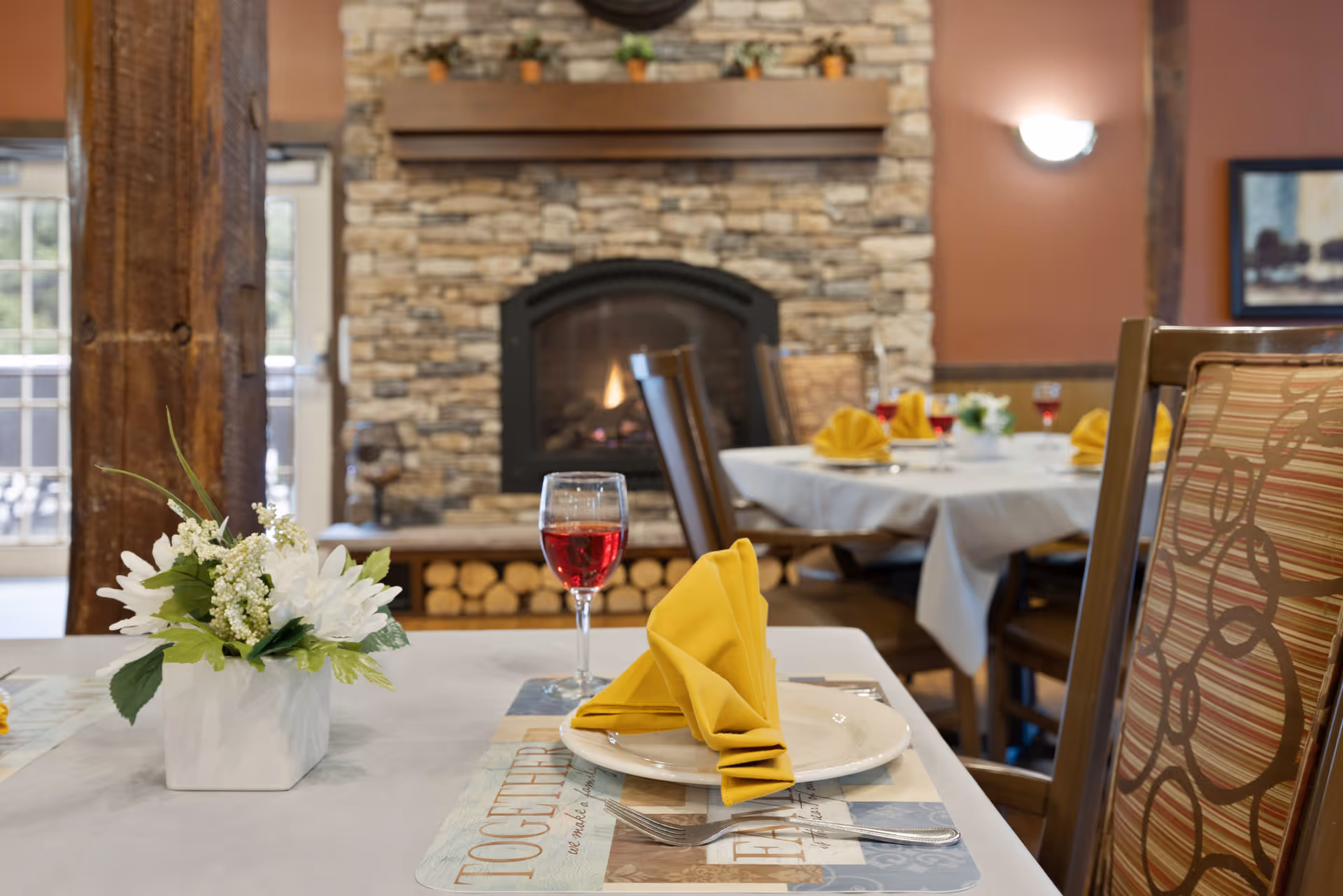 A cozy dining area with tables set for a meal, featuring white tablecloths, yellow folded napkins on white plates, glasses of red beverage, and a small floral centerpiece. In the background, there is a stone fireplace with a wooden mantel and stacked firewood underneath, along with wooden chairs and warm-colored walls.