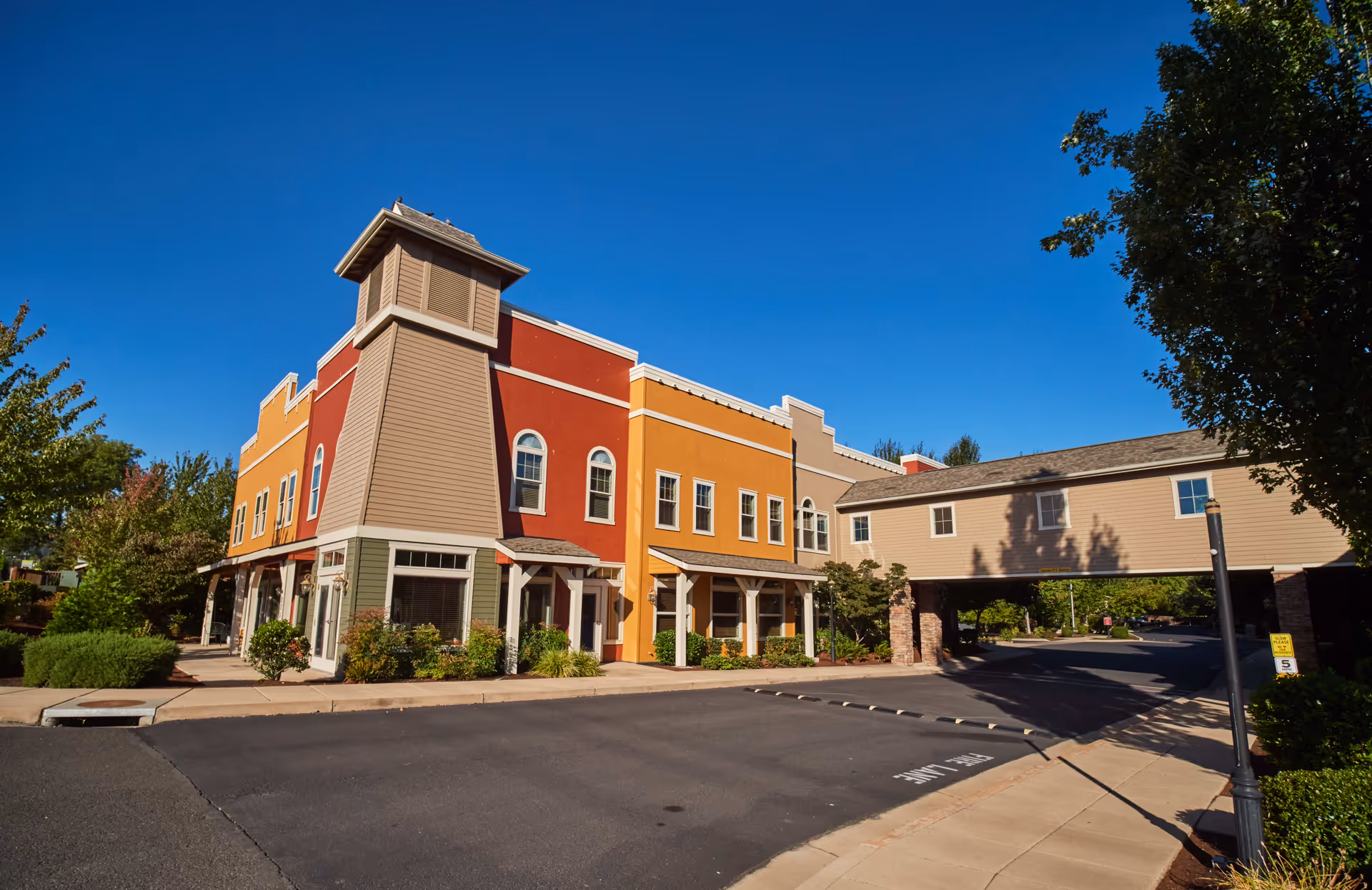 Exterior view of a colorful senior living facility building with red, orange, and beige walls under a clear blue sky. The building features a tower-like structure on the corner and a covered driveway supported by stone pillars. Trees and shrubs surround the paved driveway and sidewalk.
