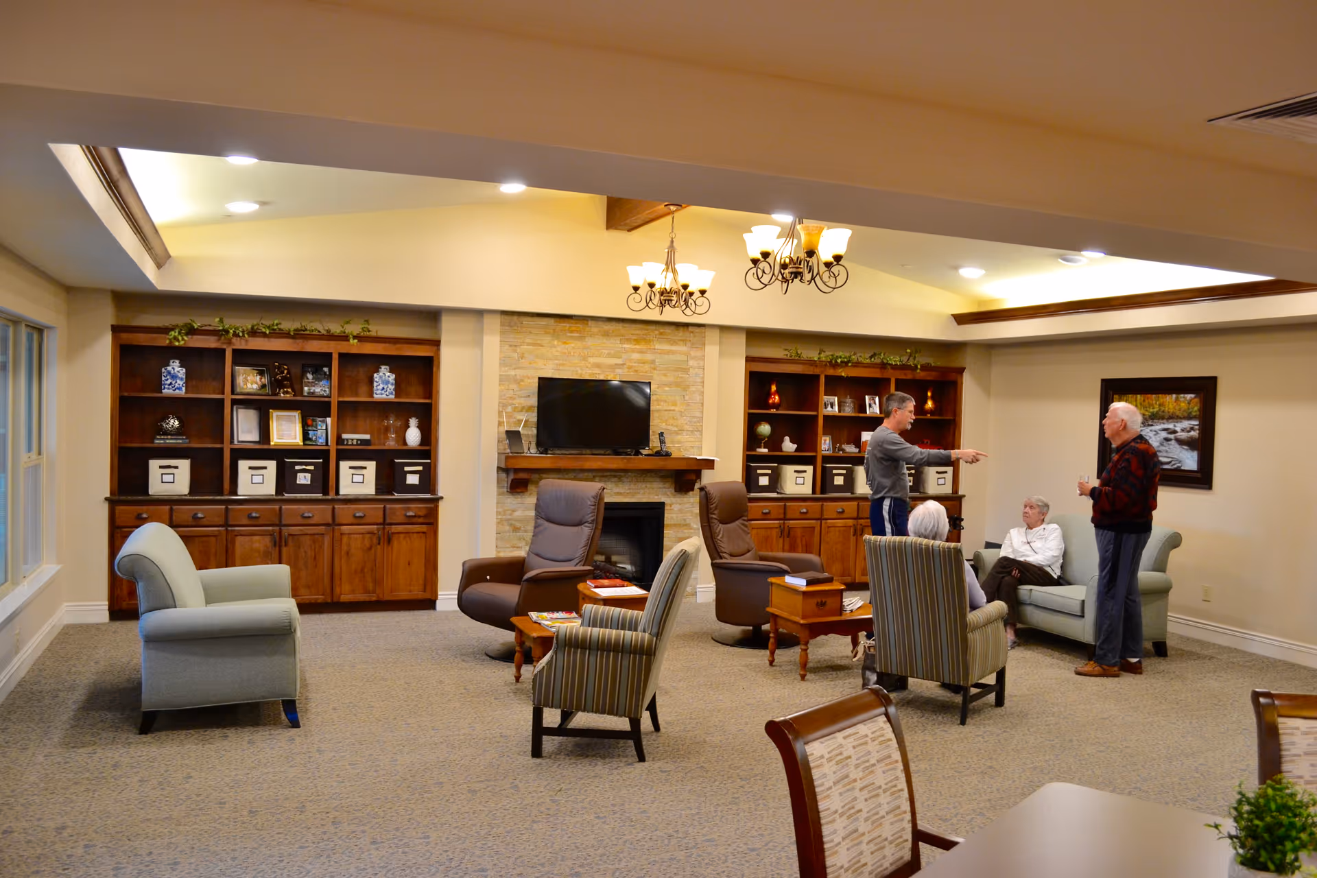 A cozy living room area in a senior living facility with several armchairs and a loveseat arranged around a fireplace with a mounted TV. There are wooden bookshelves with decorative items and storage boxes on either side of the fireplace. Four seniors are engaged in conversation, two standing and two seated. The room is warmly lit with ceiling lights and chandeliers.
