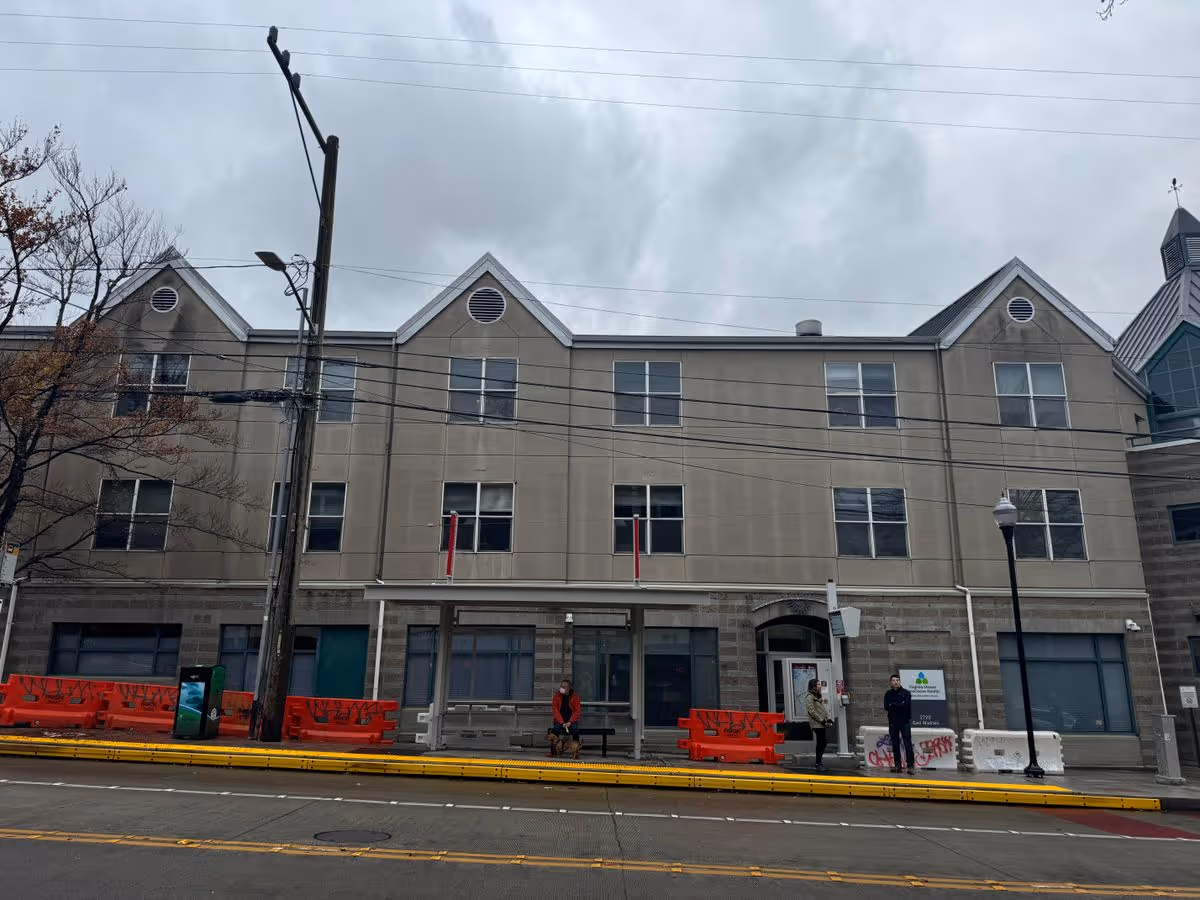Three-story beige building front with gabled roof sections, orange sidewalk barriers, and a few people near the entrance on a cloudy day.