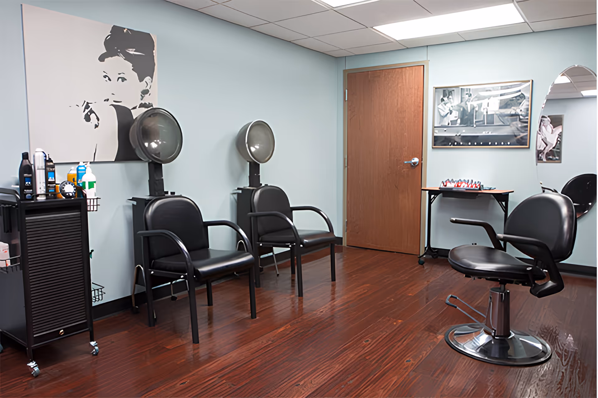 Small hair salon room with hooded dryers, black styling chairs, a barber chair, and a cart on polished wood floors.