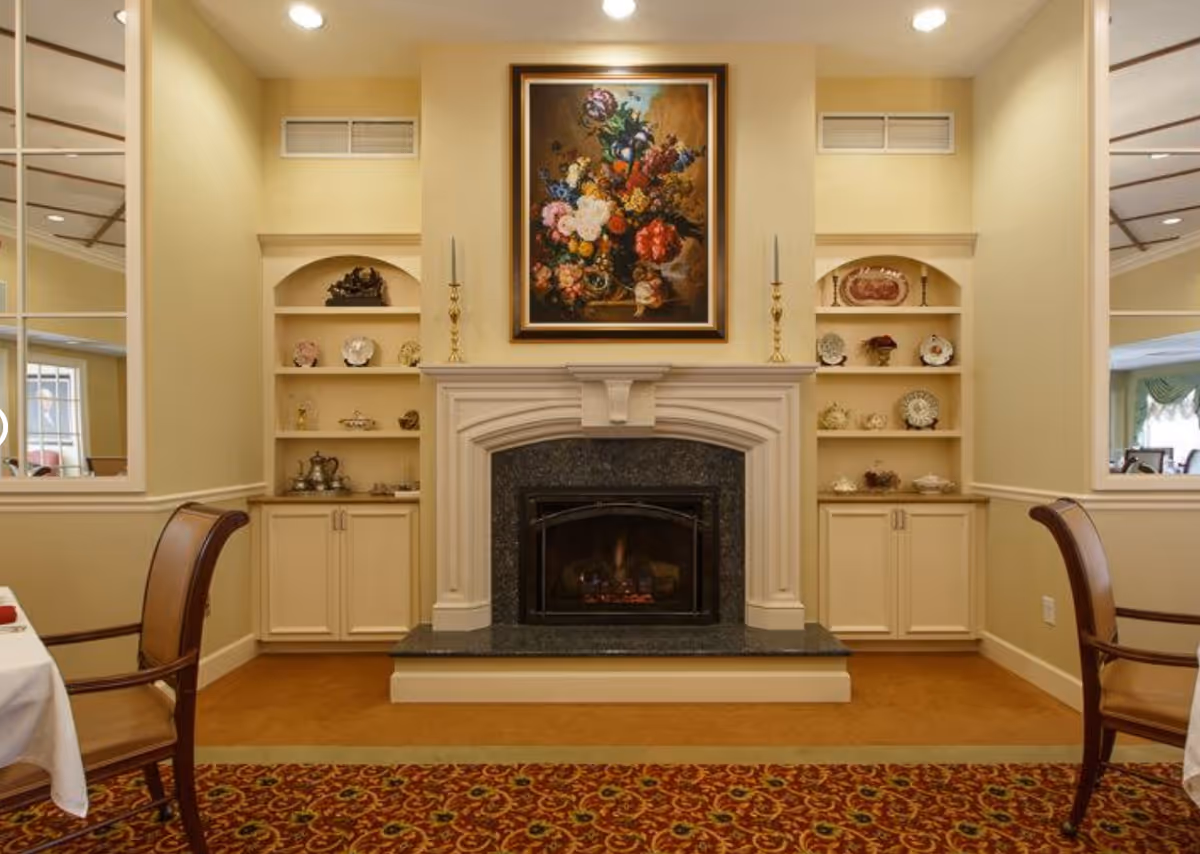 A cozy indoor space featuring a lit fireplace with a dark granite surround and white mantel. Above the fireplace hangs a framed painting of a colorful floral arrangement. On either side of the fireplace are built-in shelves displaying decorative plates, vases, and candlesticks. Two wooden chairs with cushioned seats are partially visible on each side, and the floor is covered with a patterned carpet in warm tones.