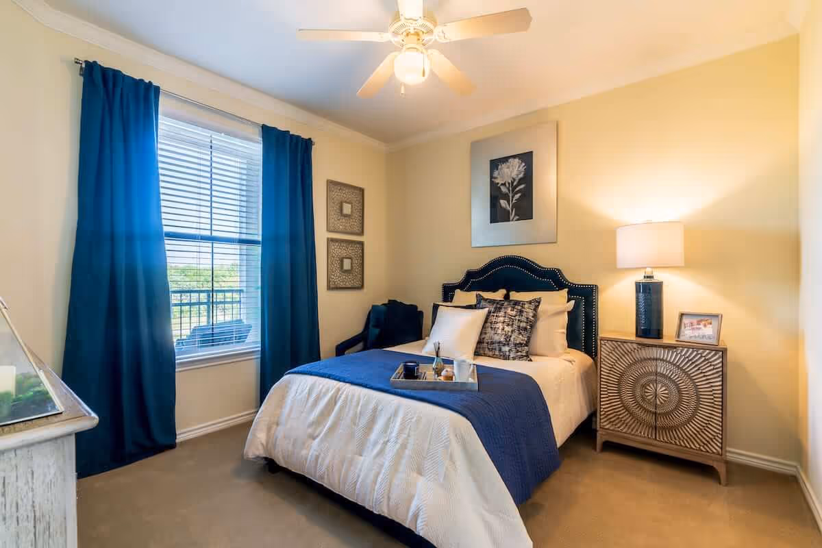 Well-lit bedroom with a bed dressed in blue and white linens, dark blue curtains at a window, a nightstand with a lamp, and wall art above the headboard.