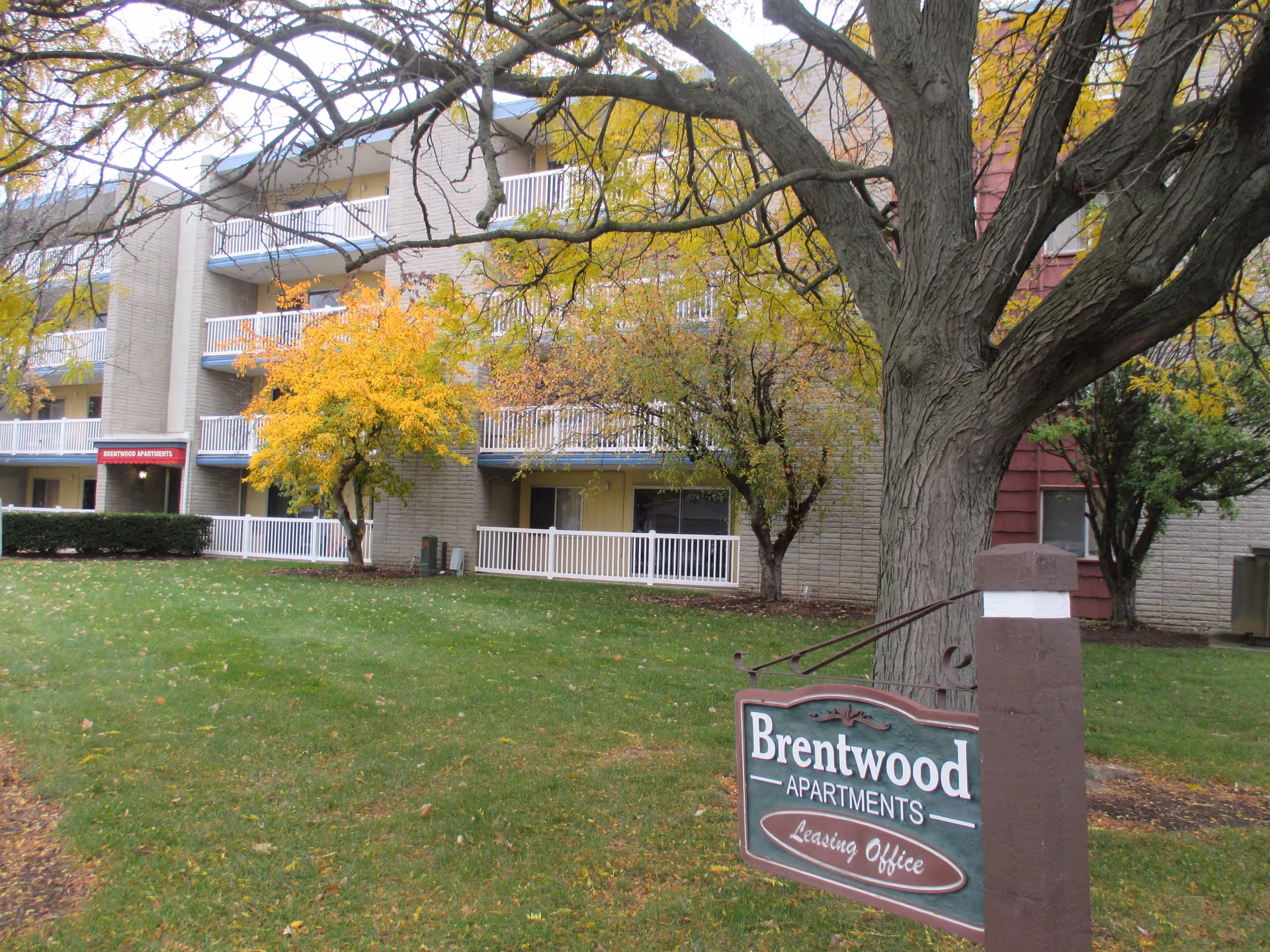 Exterior view of Brentwood Apartments building with multiple balconies, surrounded by trees with autumn foliage and a green lawn. A sign in the foreground reads 'Brentwood Apartments Leasing Office'.