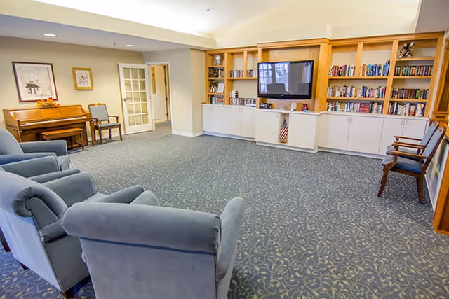 Spacious senior living common room with upholstered chairs, built-in bookshelves, a wall-mounted TV and a piano.
