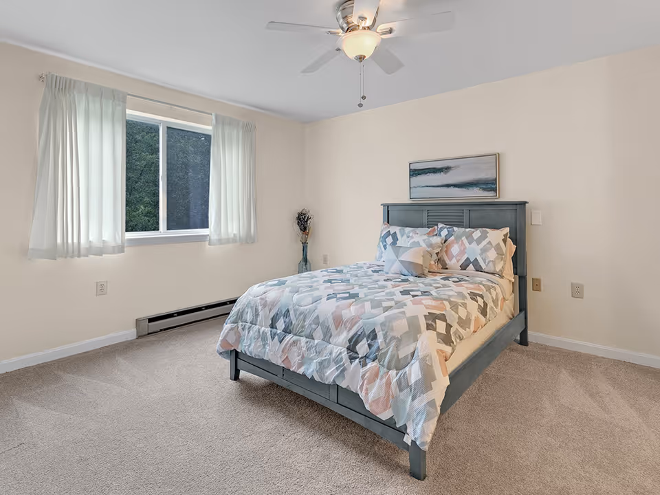 Bright bedroom with a bed in patterned linens, a window with white curtains, a ceiling fan, and neutral carpeting.